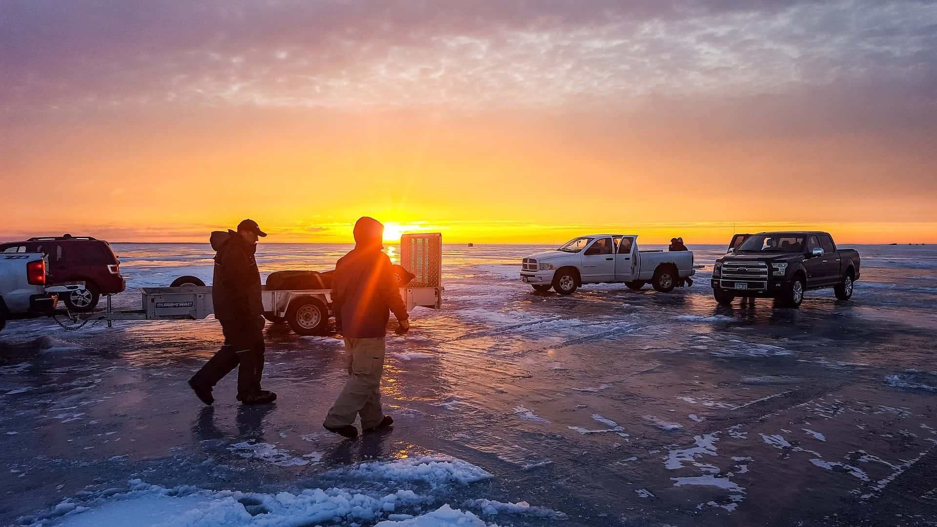 Ice Fishing 桌布 壁紙 — 兩個人在日落時分走過結冰的湖面，卡車停在冰上，天空呈現鮮豔的橙色和紫色。