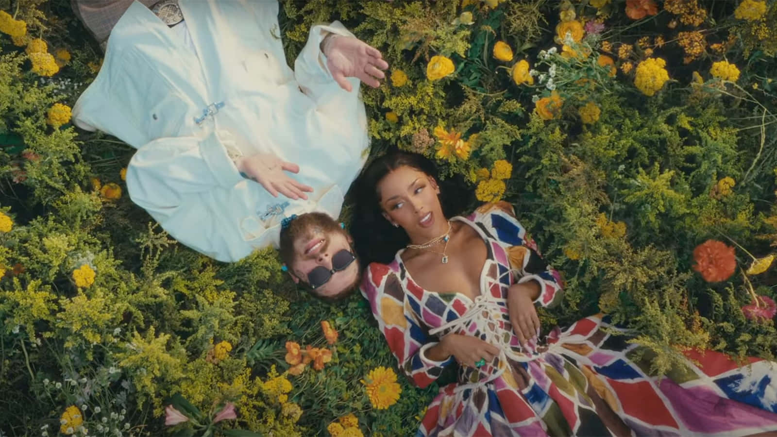 Post Malone 桌布 壁紙 — 兩個人在明亮的日光下，放鬆地躺在充滿黃色和橙色野花的生機勃勃的田野中。