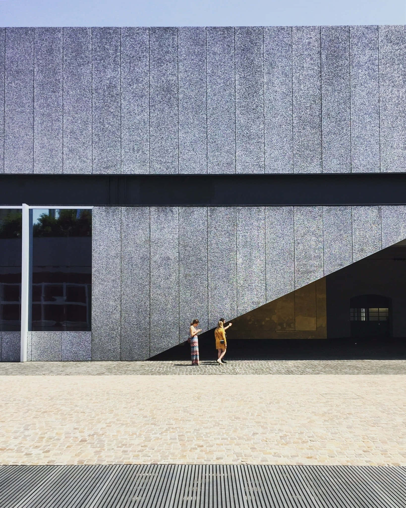 Modernism 桌布 壁紙 — 兩個人在明亮的日光下站在一座巨大的紋理灰色混凝土建築前，投射出清晰的陰影。