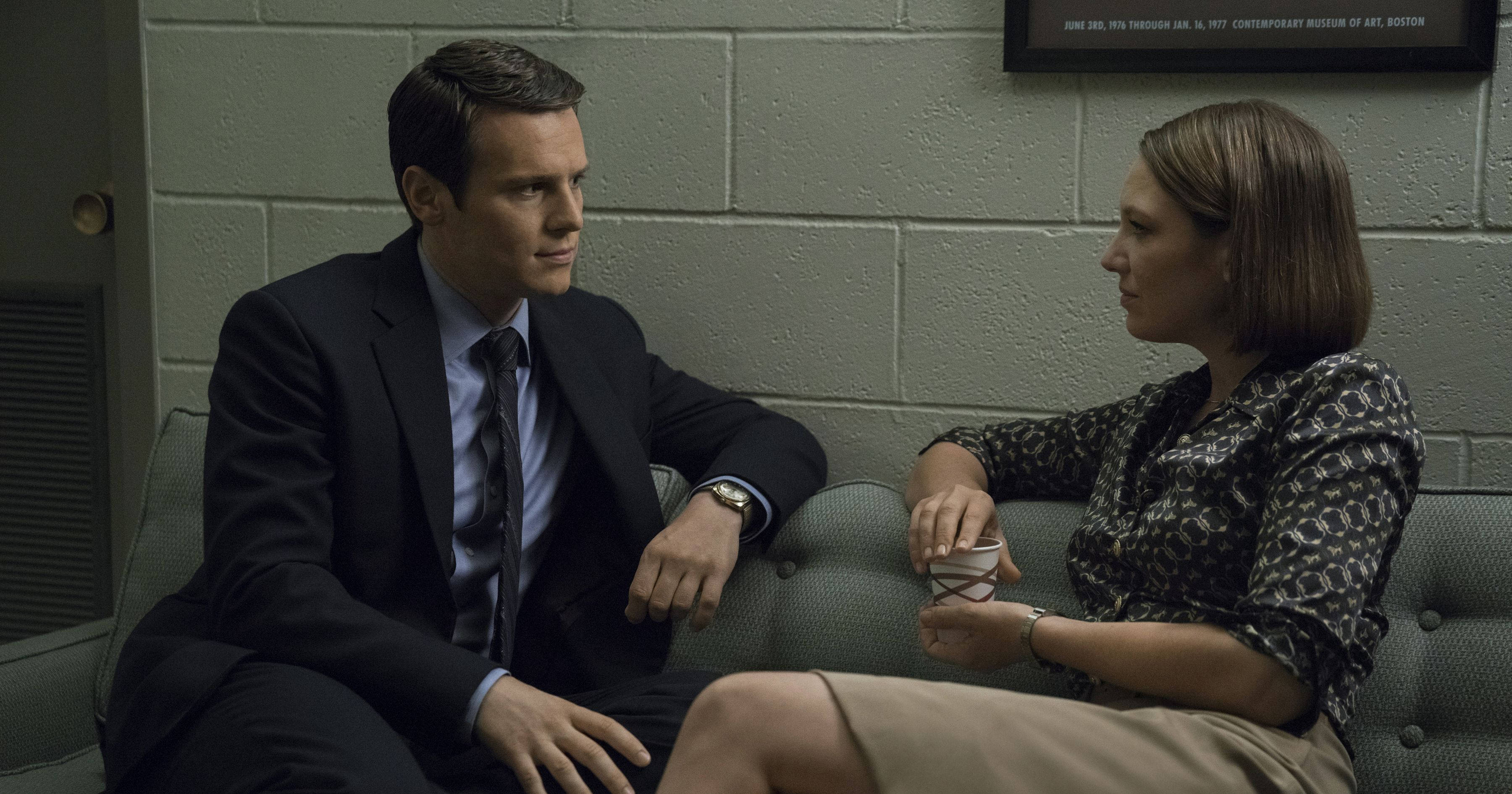 Jonathan Groff And Anna Torv Mindhunter Stills 壁紙 — 兩個人坐在灰色梳化上，身處一個只有煤渣磚牆的簡陋房間，正在進行嚴肅對話。