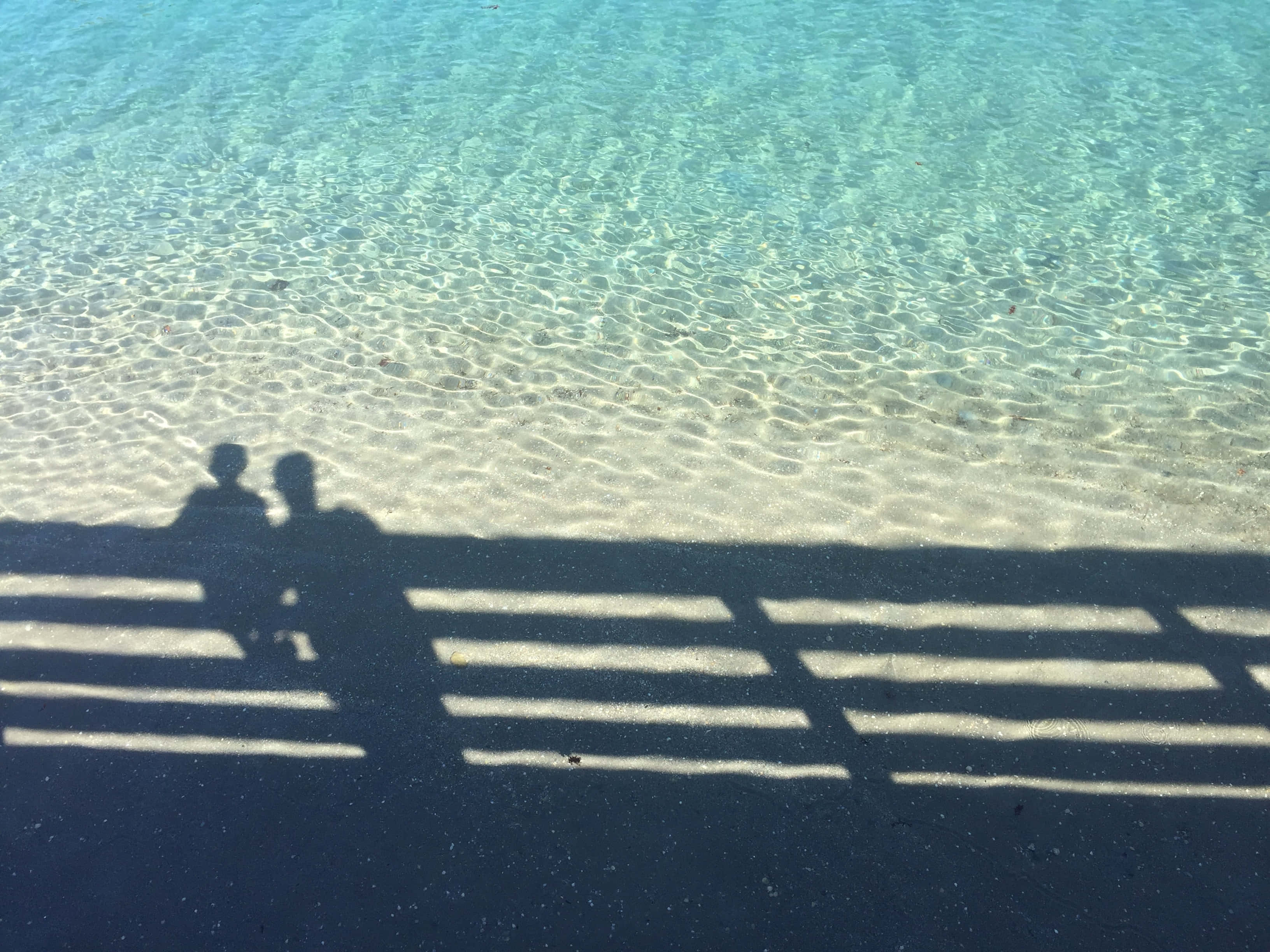 Summer Vacation 桌布 壁紙 — 兩個人坐在長椅上的剪影，在漣漪碧綠海水旁的深色沙灘上投下長長的影子。
