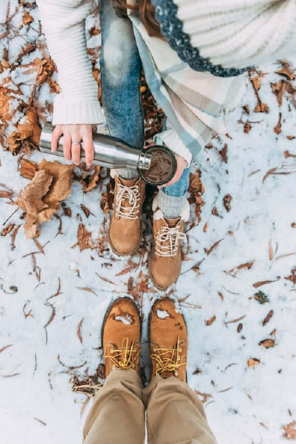 Snowshoes 桌布 壁紙 — 兩個人坐在雪地上，周圍有落葉，其中一人穿著靴子，從保溫壺倒咖啡到杯子裡