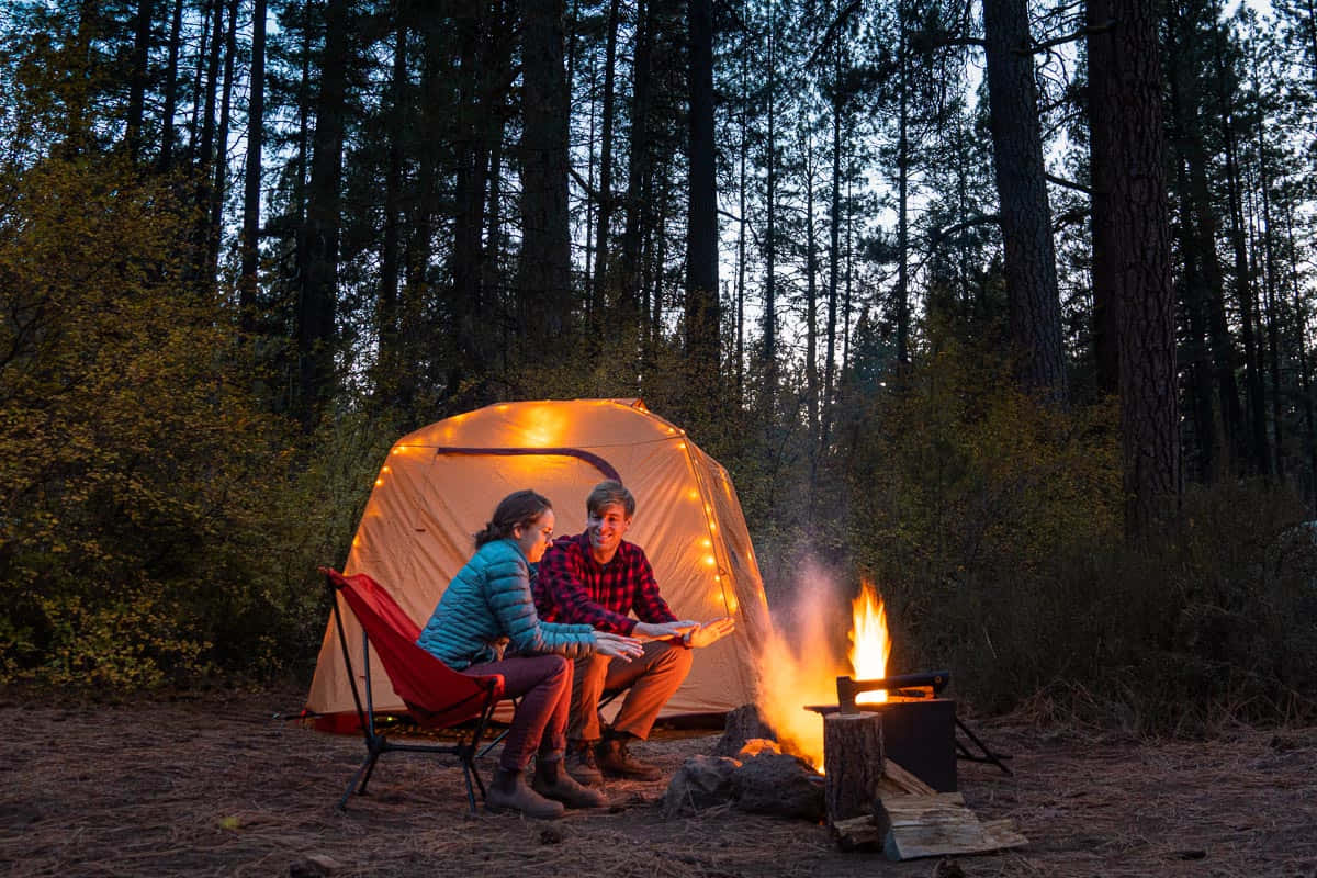 Fall Camping 桌布 壁紙 — 兩個人坐在黃昏的營火旁，在松樹林中發光的帳篷旁邊看書。