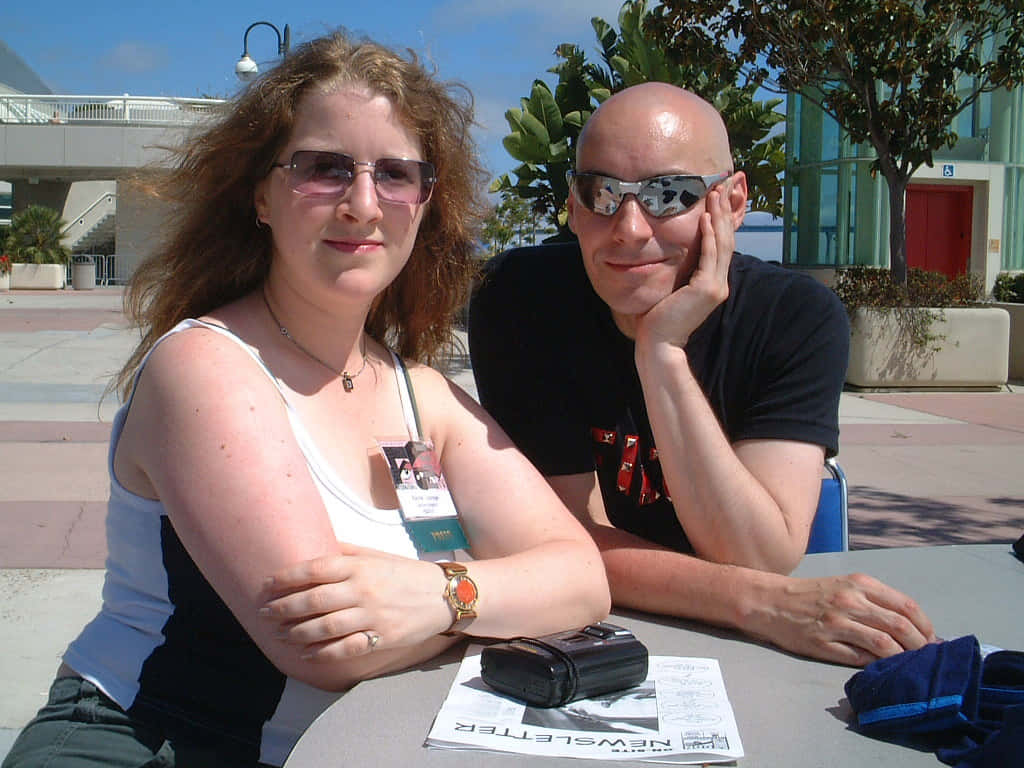 Grant Morrison 桌布 壁紙 — 兩個人戴著太陽眼鏡坐在戶外餐桌，背景有陽光、現代建築和綠化。