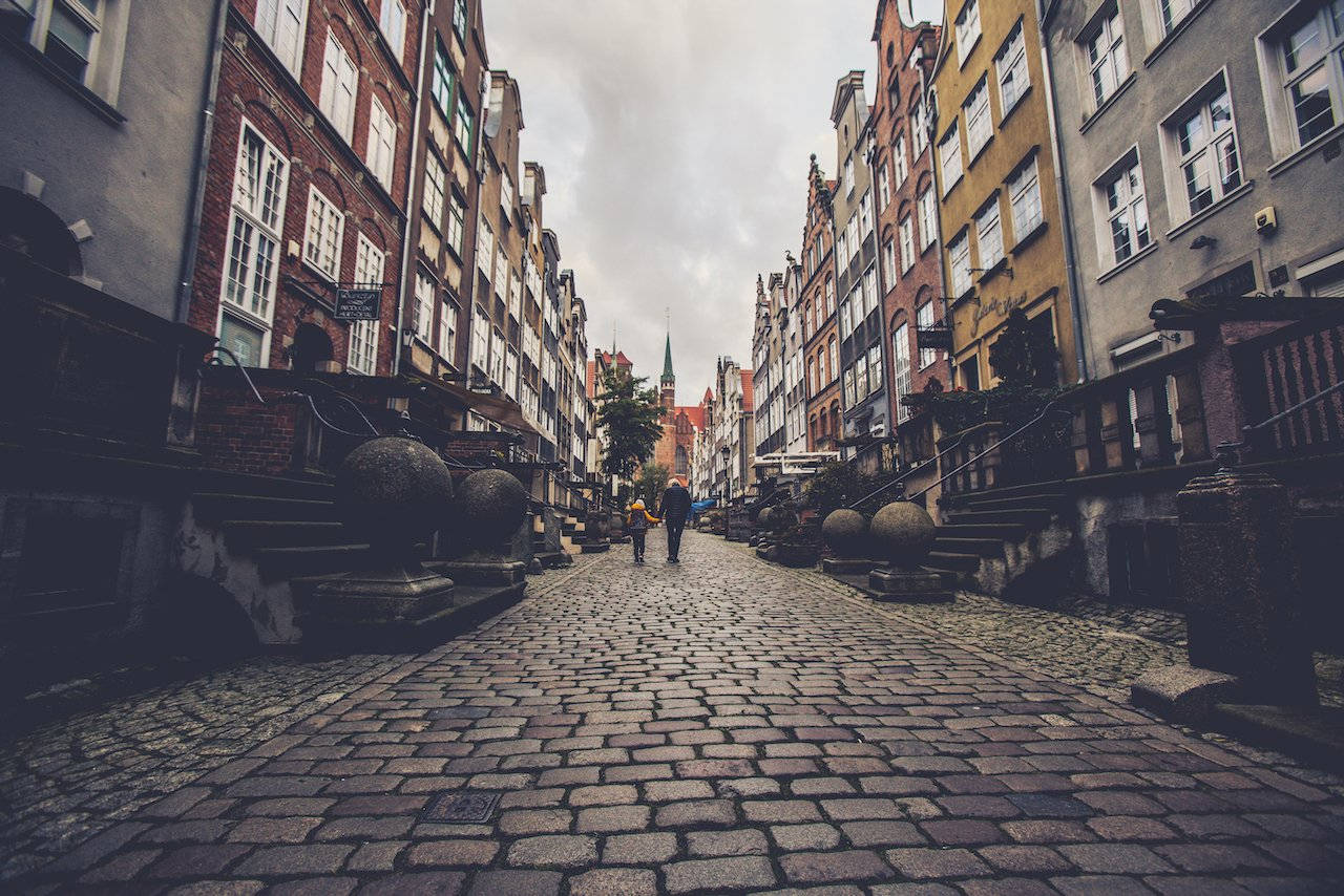 Crooked House 桌布 壁紙 — 兩個人物喺陰天嘅天空下，沿住一條狹窄嘅鵝卵石街道散步，兩旁係歷史悠久嘅歐洲建築。