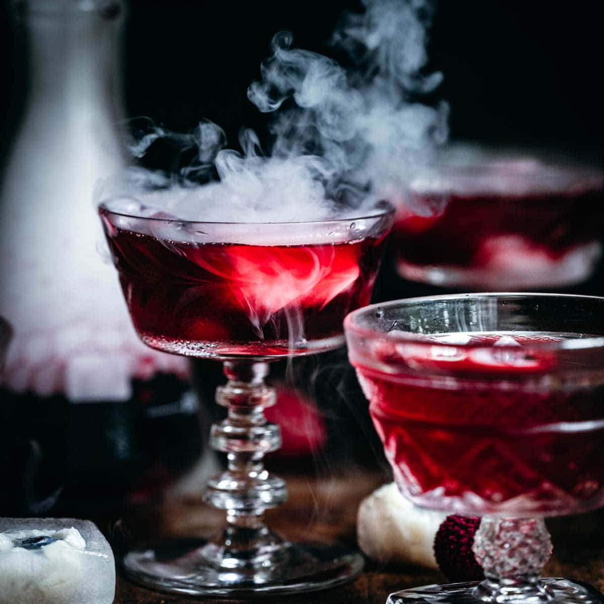 Halloween Cocktails 桌布 壁紙 — 兩個優雅的玻璃杯裝滿發光的紅色雞尾酒，在黑暗背景下散發出旋轉的煙霧。