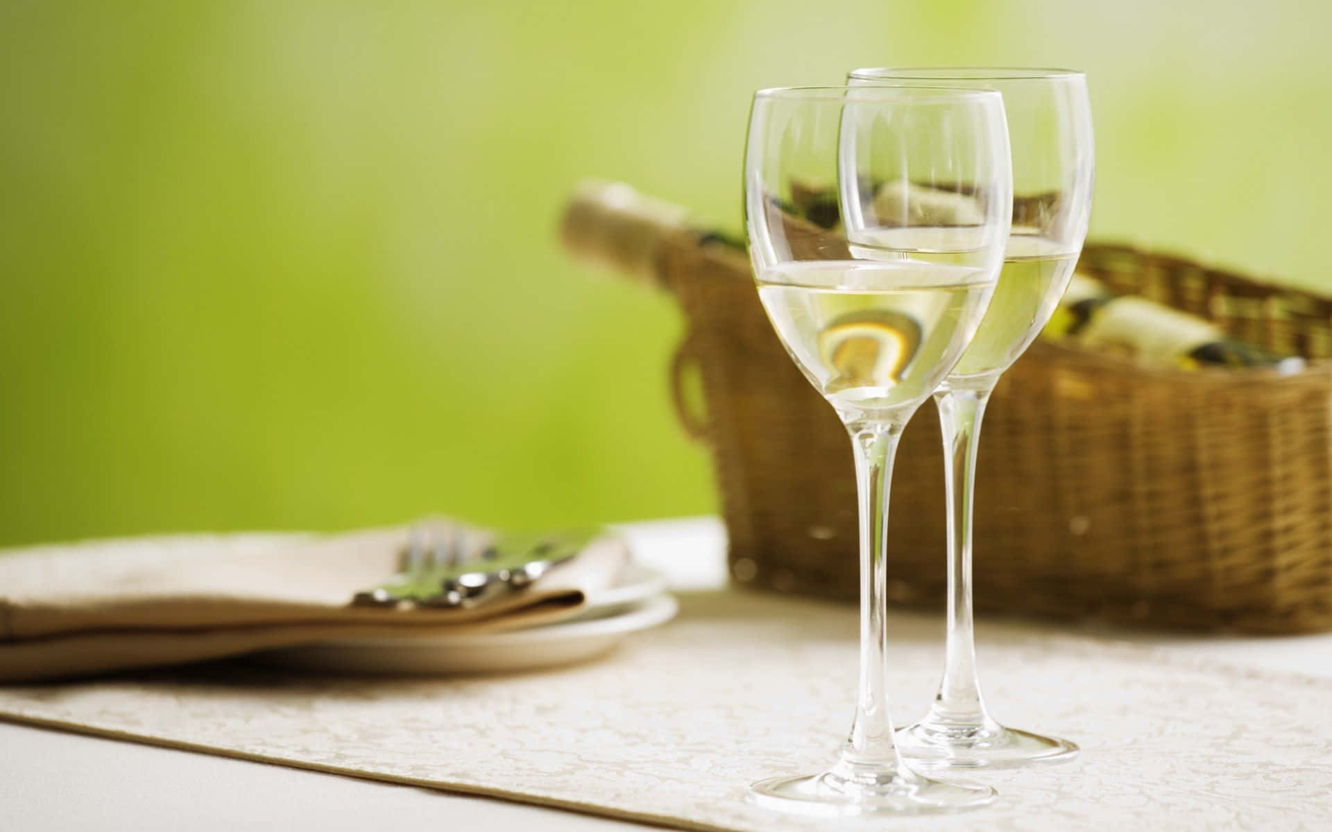 White Wine 桌布 壁紙 — 兩個優雅的酒杯盛滿白酒，放在蕾絲桌布上，旁邊有一個柳條野餐籃，背景是柔和的綠色。