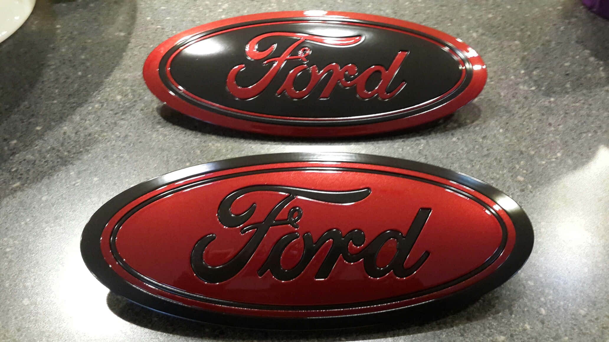 Ford Logo 桌布 壁紙 — 兩個光面福特標誌，一個紅色配黑色字，另一個黑色配紅色字，放在有斑點的灰色枱面上。