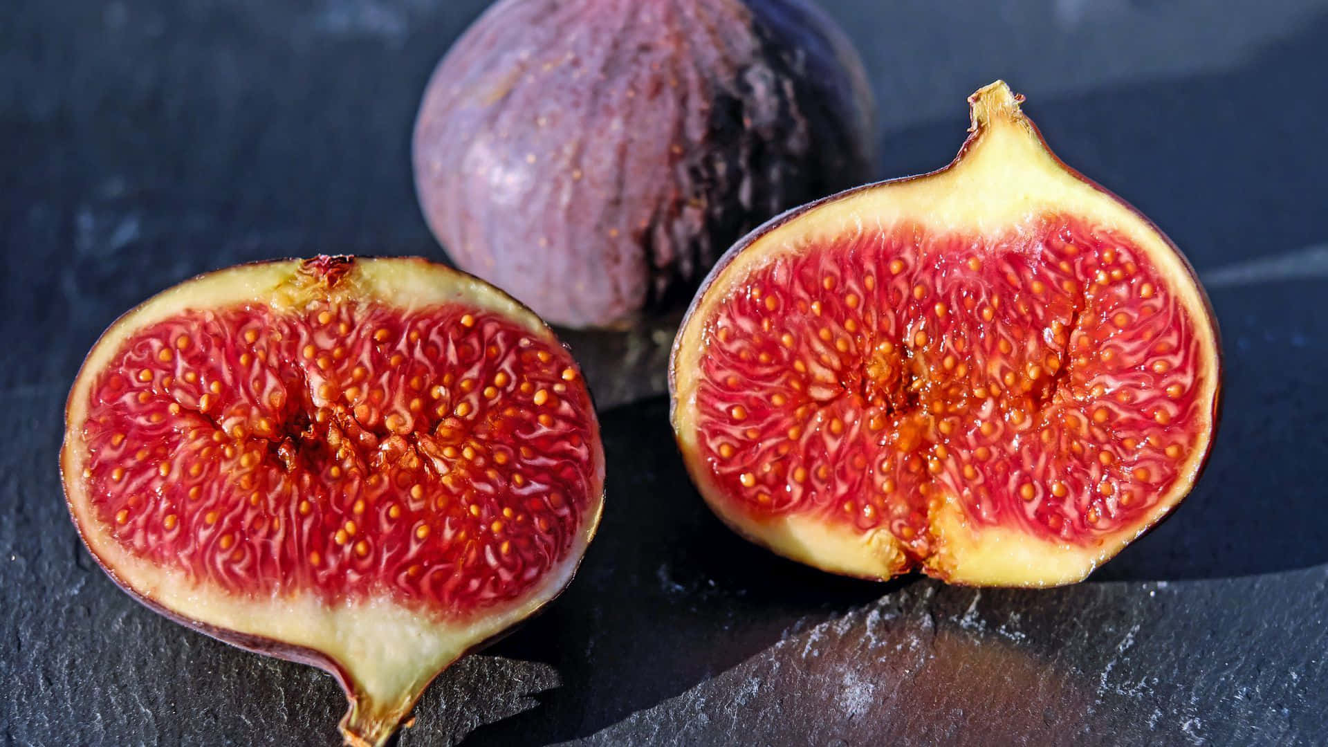 Purple Figs 桌布 壁紙 — 兩個切半的成熟無花果，露出多汁的紅色果肉和細小種子，與一個完整的無花果一起放在深色石板之上。