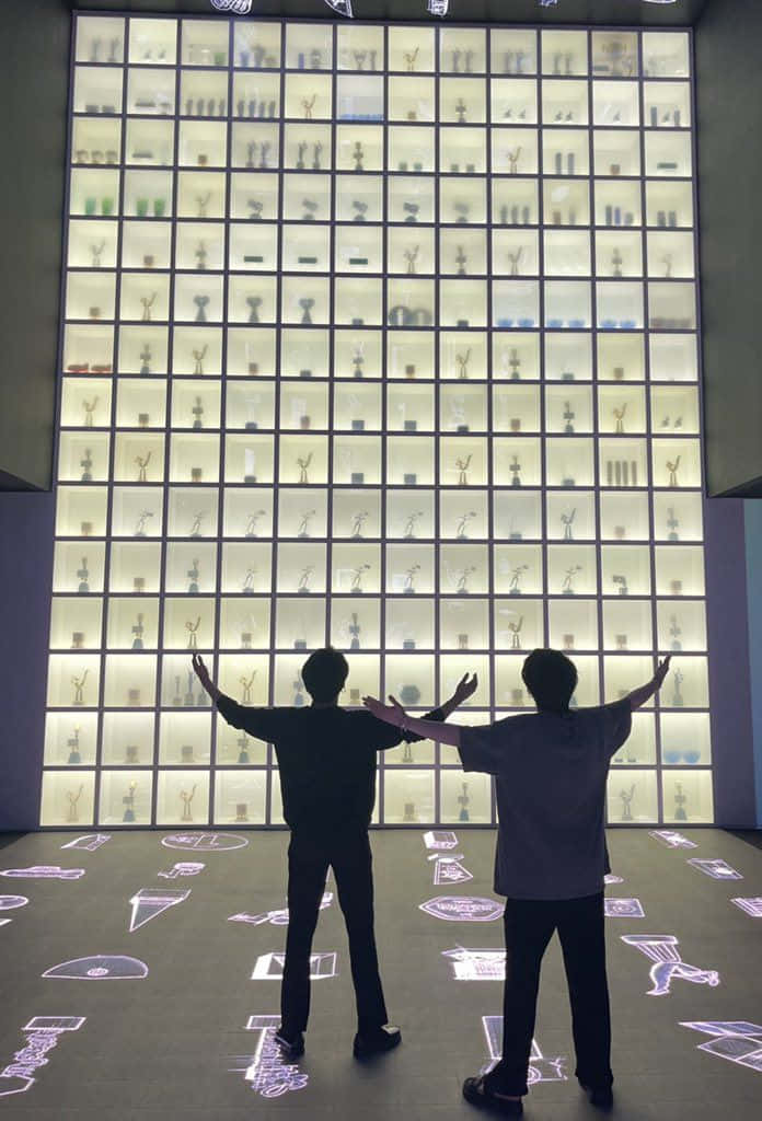 Bts Endorsements 桌布 壁紙 — 兩個剪影人物在一個發光的網格架子前伸展雙臂，架子上擺滿了微型獎盃和工藝品。