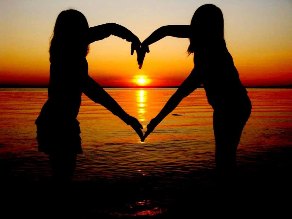 Heart Silhouette 桌布 壁紙 — 兩個剪影人物在日落時分於平靜的水面上用手形成一個心形，水面倒映著發光的天空。