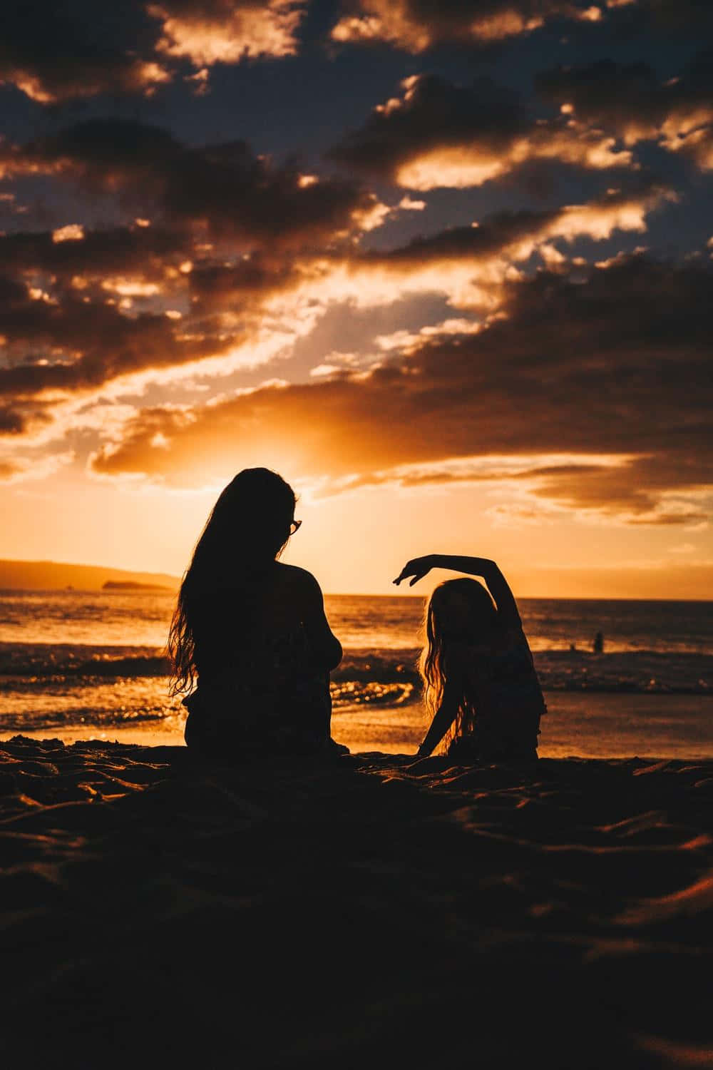 Sunset Silhouette 桌布 壁紙 — 兩個剪影人物坐在沙灘上欣賞日落，看著海浪，上方熾熱的橙色雲朵閃耀著光芒。