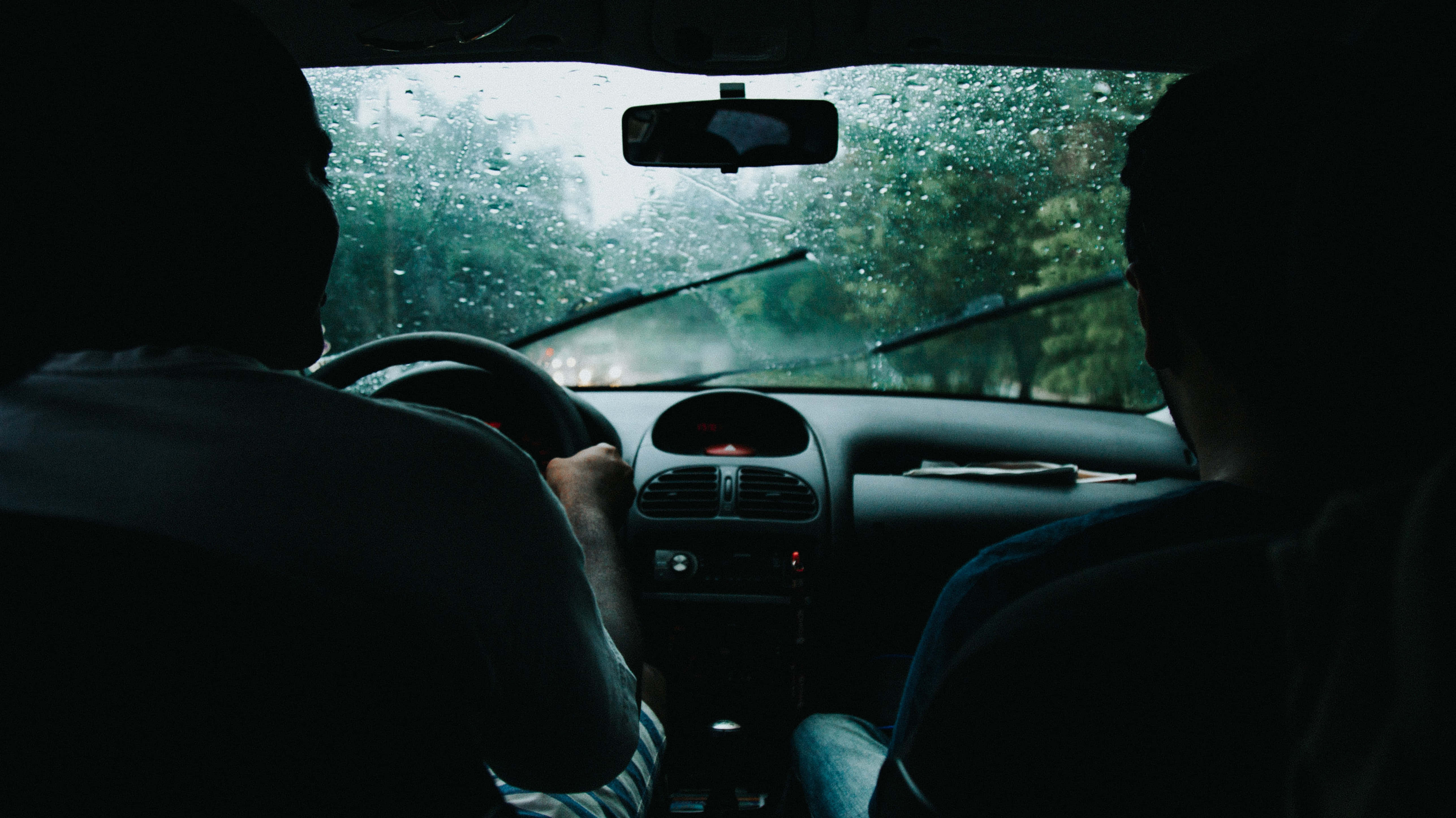 road trip 桌布 壁紙 — 兩個剪影人物坐在車內，雨水在擋風玻璃上流淌，從後座以昏暗的車內燈光觀看。
