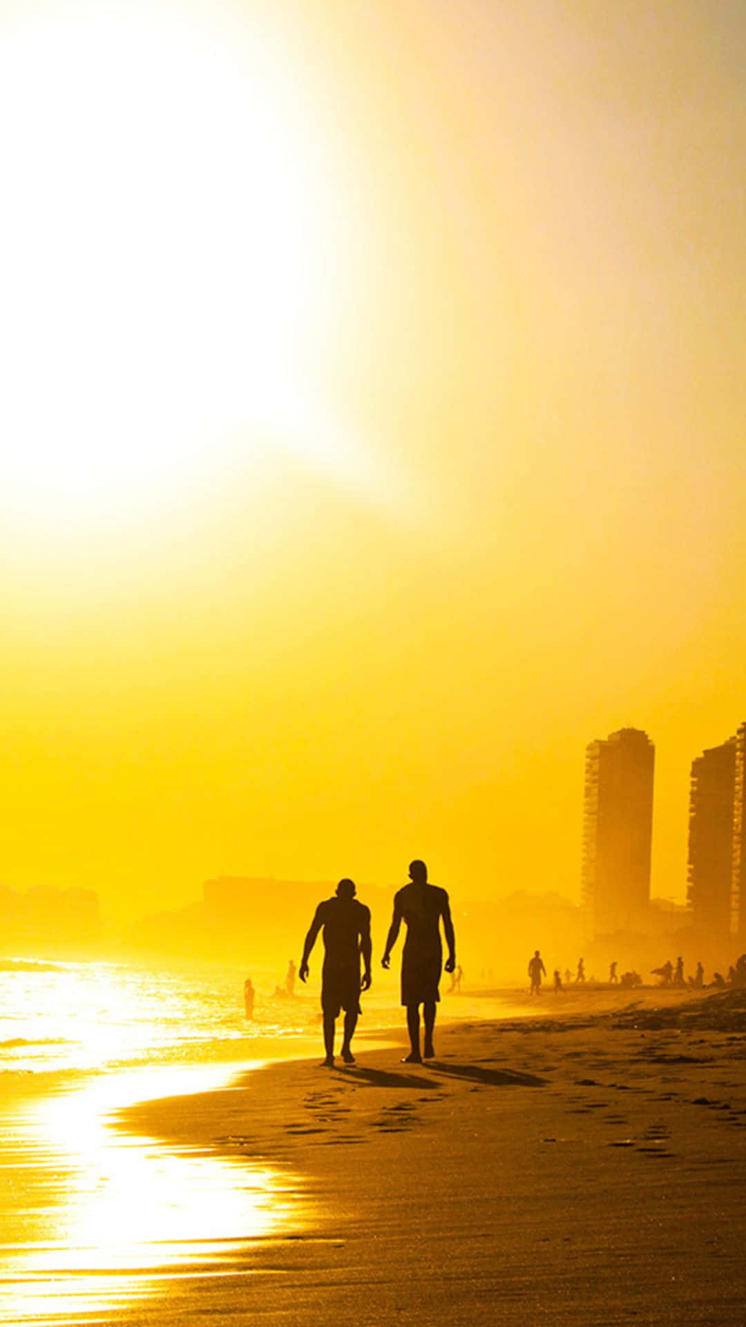 Yellow Beach 桌布 壁紙 — 兩個剪影人物沿著陽光普照的海灘漫步，金色光芒沐浴著水面和遠處的城市天際線。