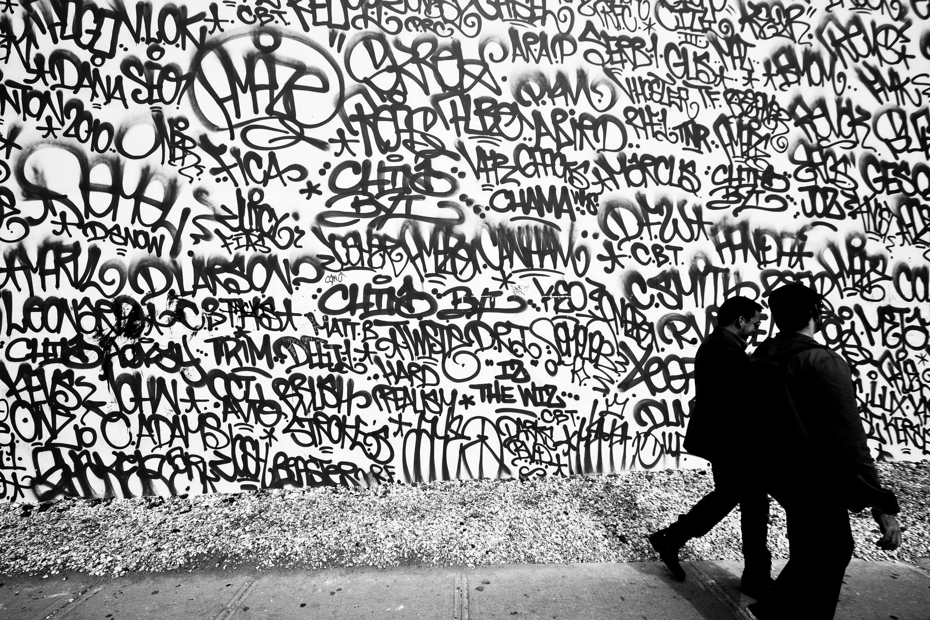 Black And White Graffiti 桌布 壁紙 — 兩個剪影人物走過一面佈滿密集混亂塗鴉的牆壁，呈現鮮明的黑白對比。