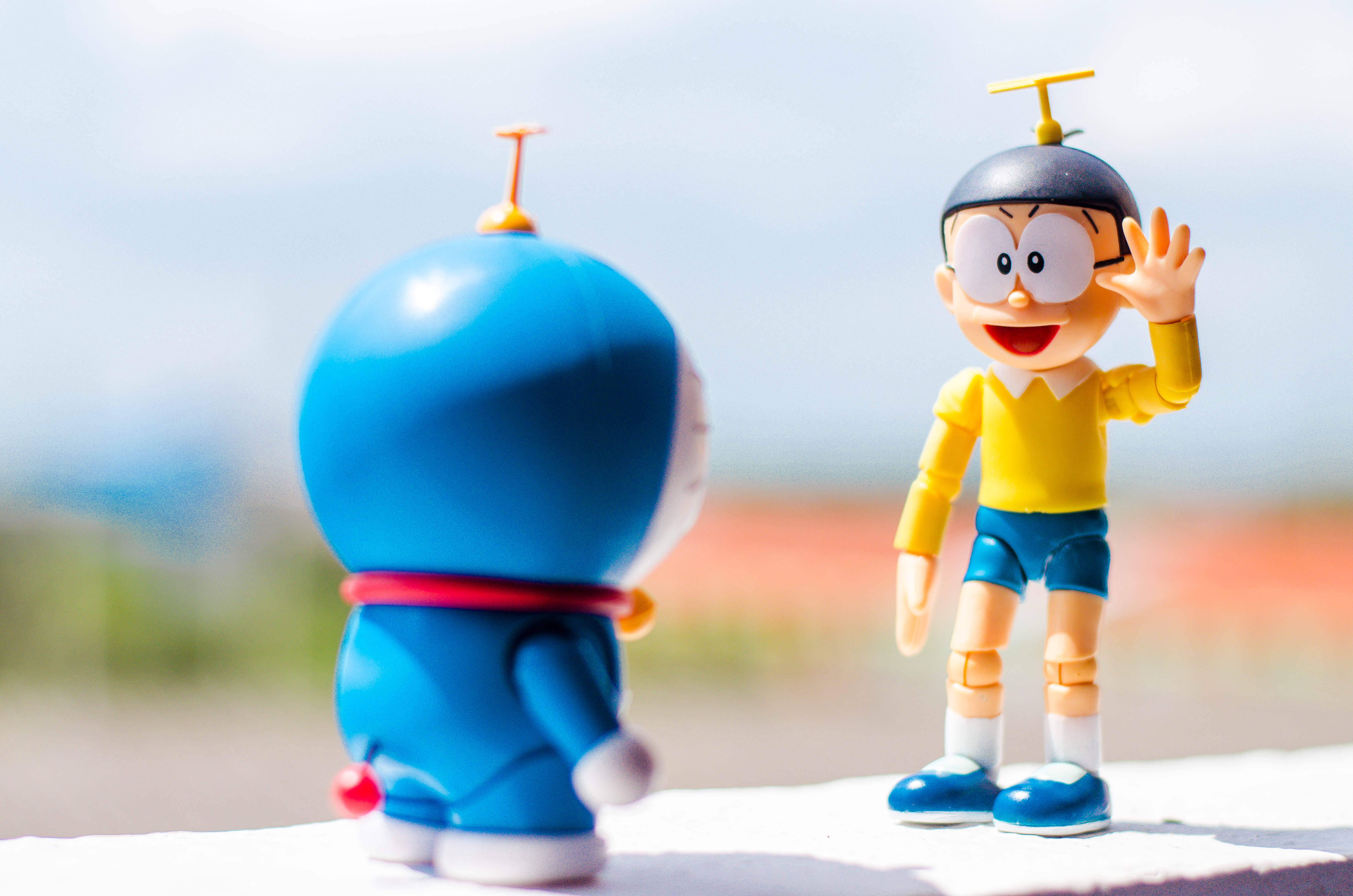 Doraemon 桌布 壁紙 — 兩個動漫人物多啦A夢和大雄在明亮的日光下，站在一個壁架上，面對面，背景是模糊的戶外景色。