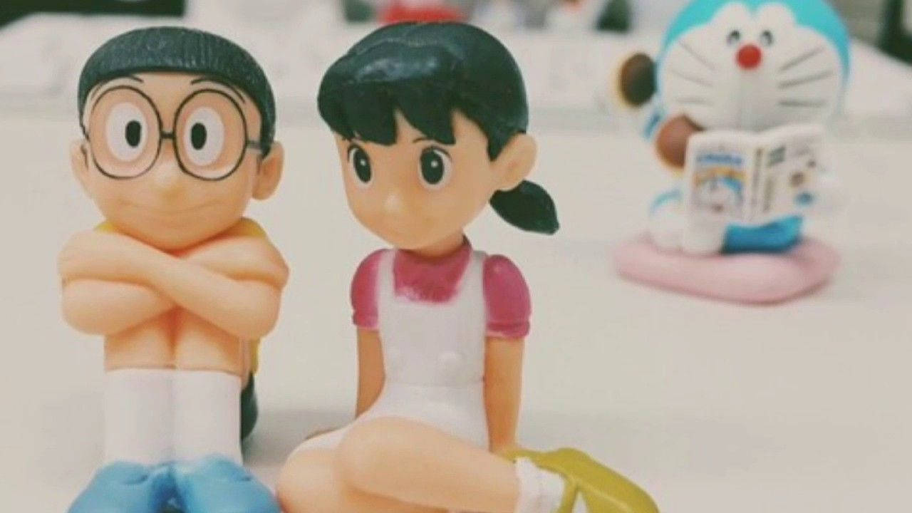 nobita shizuka love 桌布 壁紙 — 兩個動漫風格嘅公仔，一個男仔同一個女仔坐得好近，背景有一隻藍色機械貓。