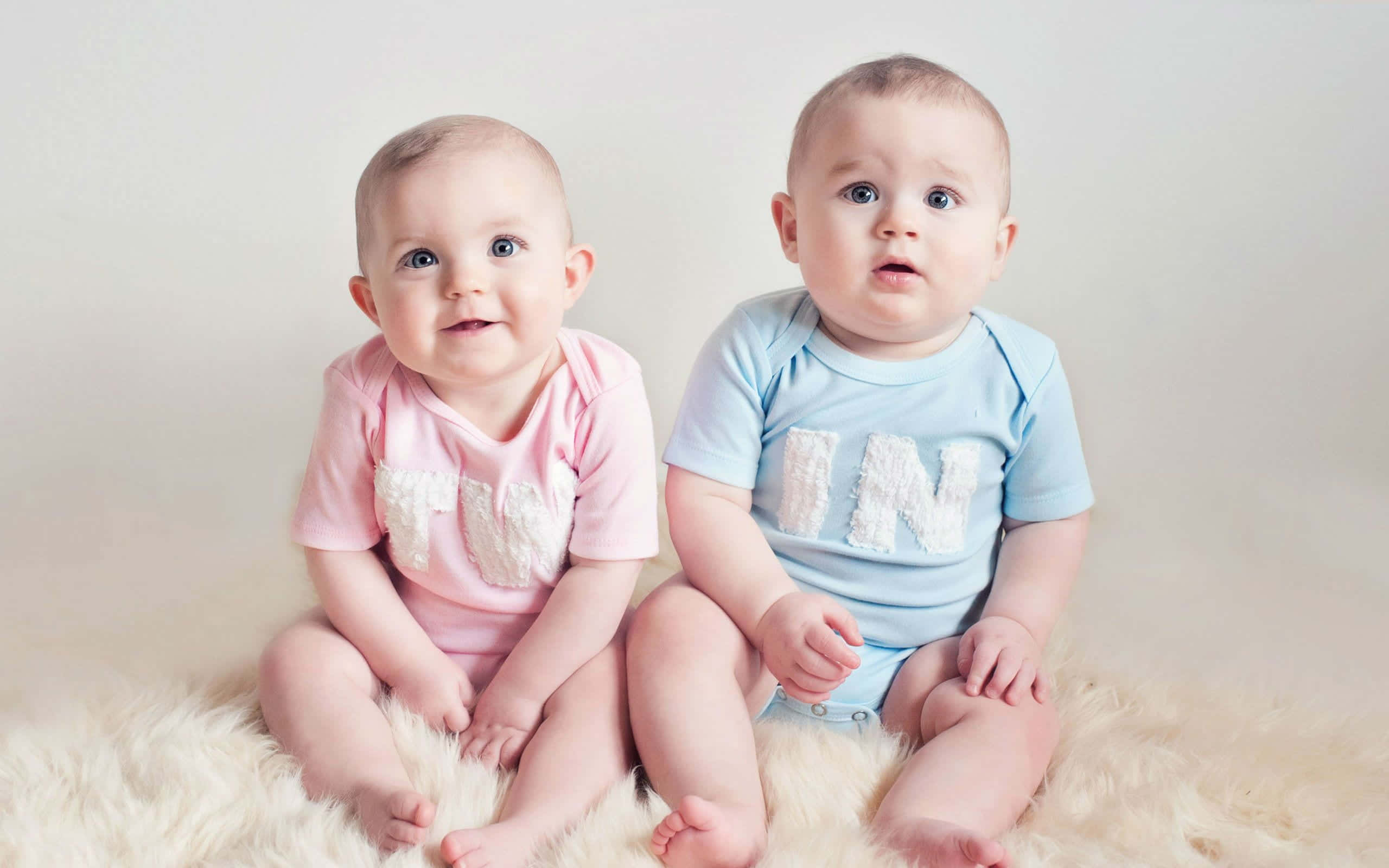 Cute Babies 桌布 壁紙 — 兩個可愛嘅BB並排坐喺一張毛茸茸嘅白色地氈上，一個着粉紅色，另一個着藍色，兩個都好奇咁望住鏡頭。