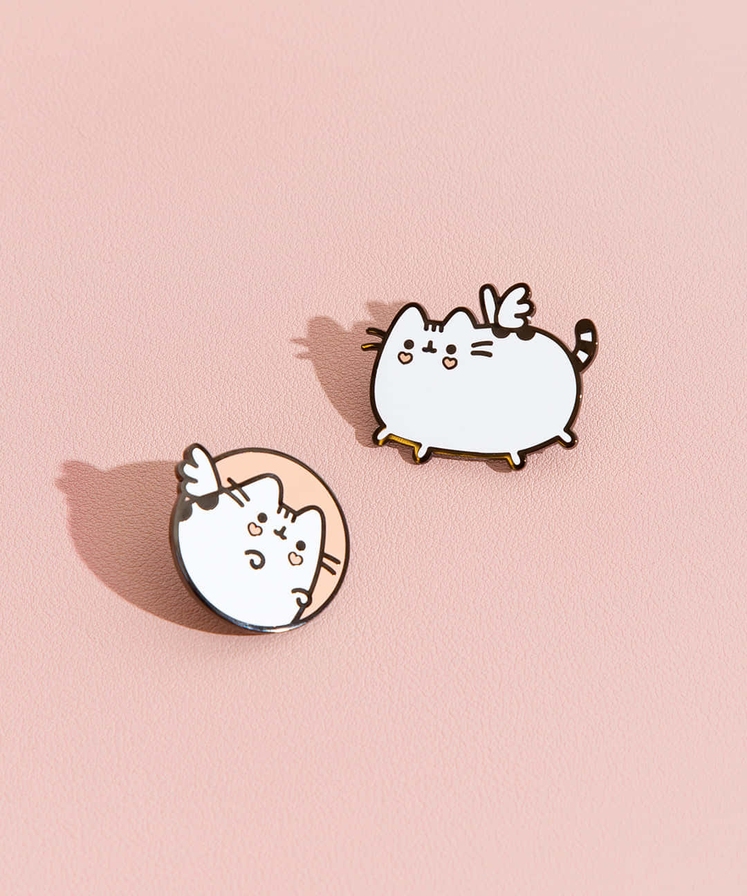 Pusheen 桌布 壁紙 — 兩個可愛的Pusheen琺瑯別針，帶有天使翅膀，放在柔軟的粉紅色紋理表面上，投下微妙的陰影。