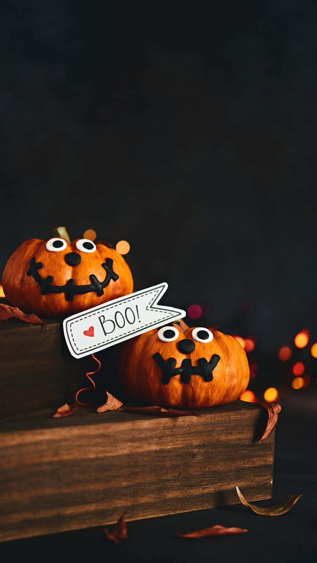 Cute Pumpkin 桌布 壁紙 — 兩個可愛的雕刻南瓜，有凸眼和縫合的嘴巴，坐在一個木箱上，上面有 'BOO!' 標誌，背景是黑暗的，帶有溫暖的散景燈光。
