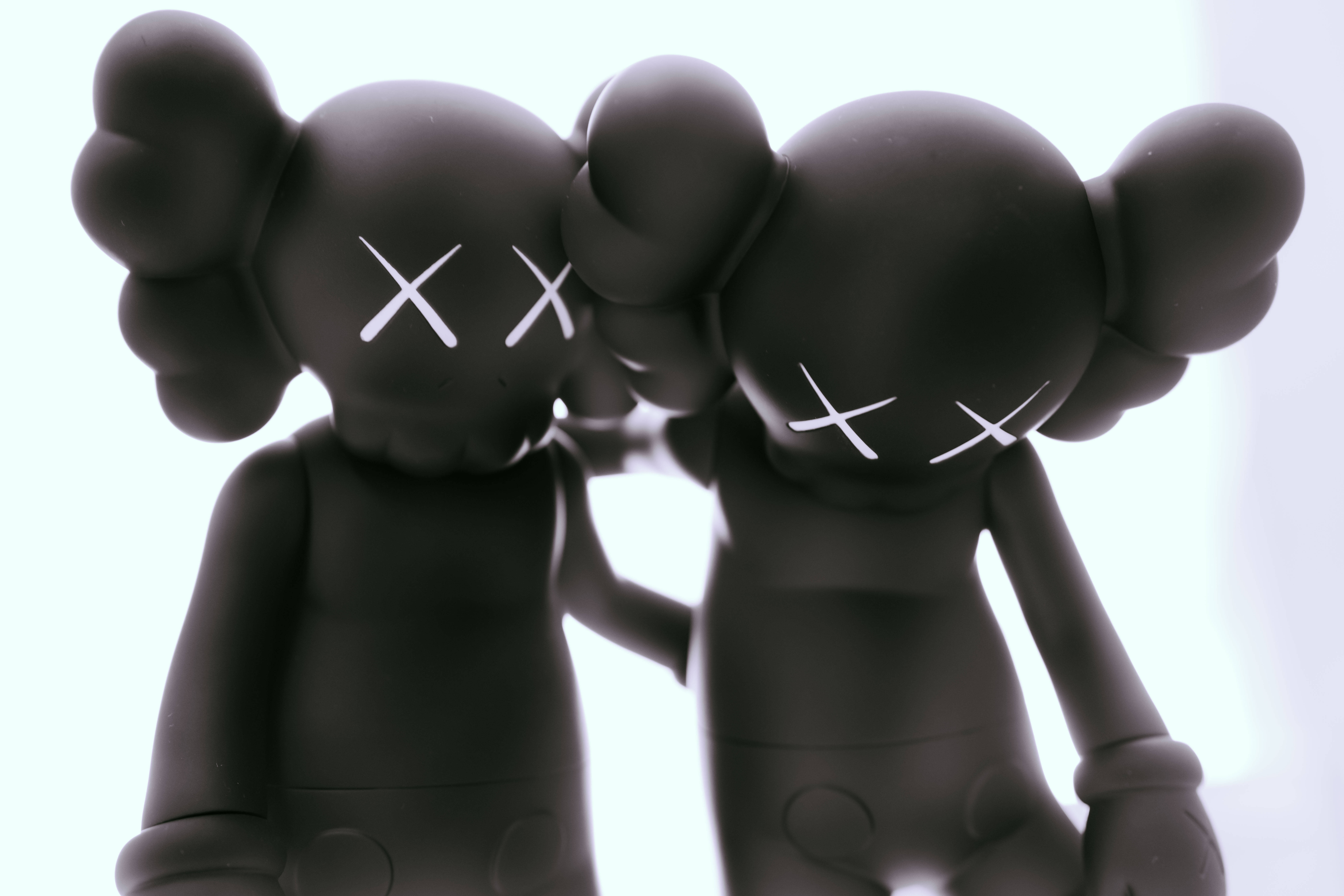 Kaws Black Along The Way 壁紙 — 兩個啞光黑色KAWS人偶，帶有白色X眼睛，在明亮的白色背景上擁抱。