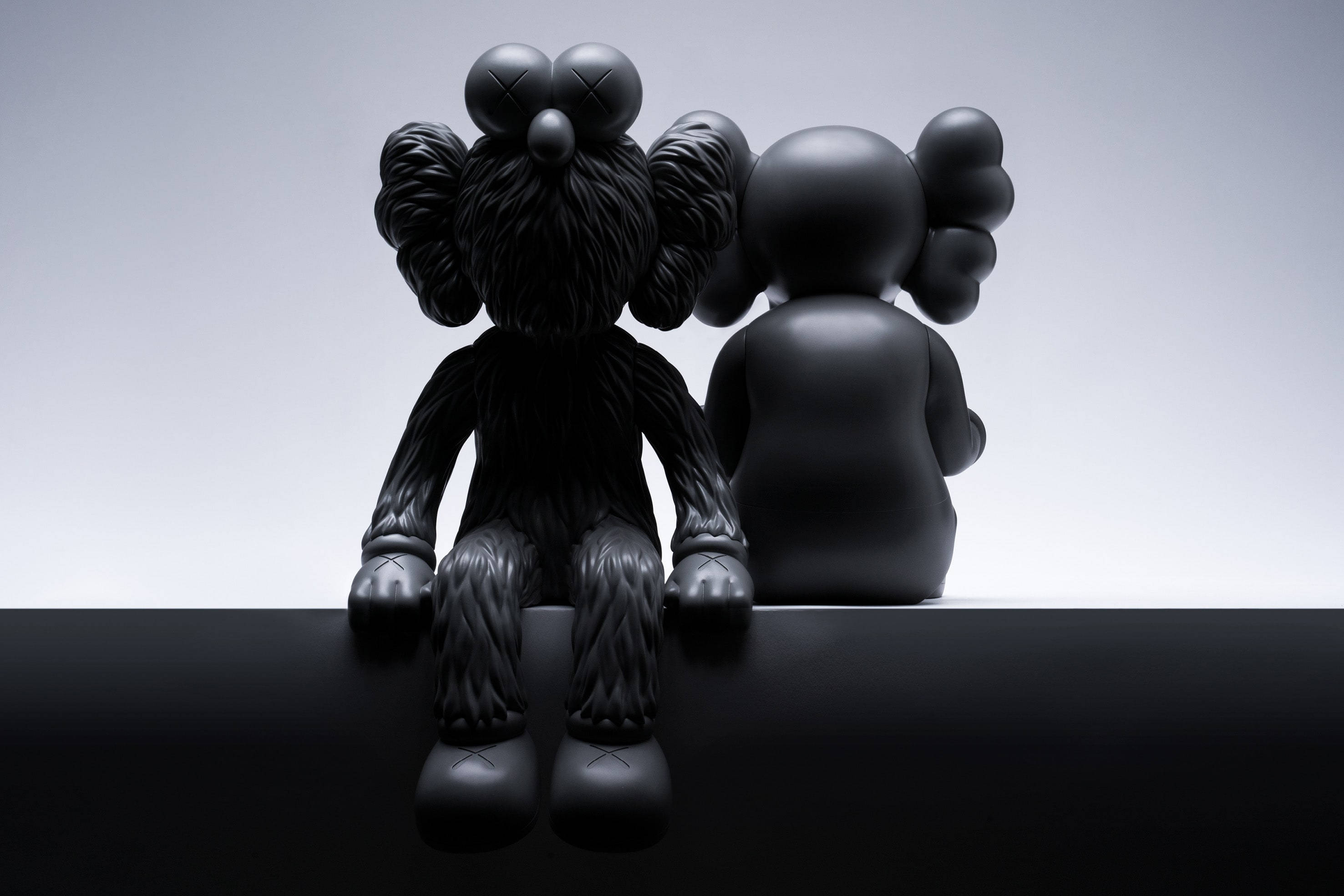 KAWS Seeing/Watching 壁紙 — 兩個啞光黑色KAWS公仔並排坐在光滑的黑色表面上，背景為漸變灰色。