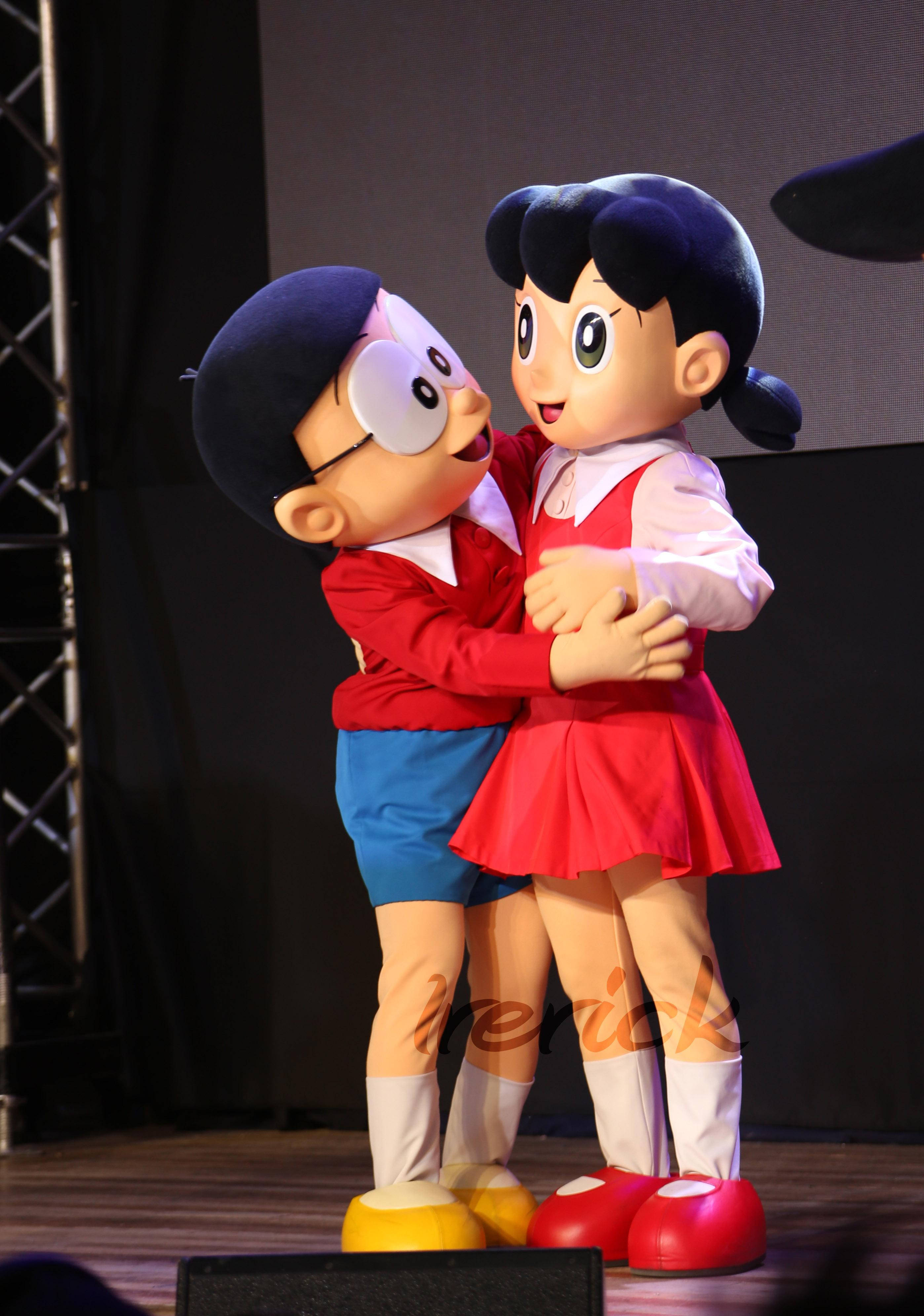 Doraemon 桌布 壁紙 — 兩個大型動漫吉祥物多啦A夢和靜香在黑暗背景和可見燈光設備的舞台上溫暖擁抱。