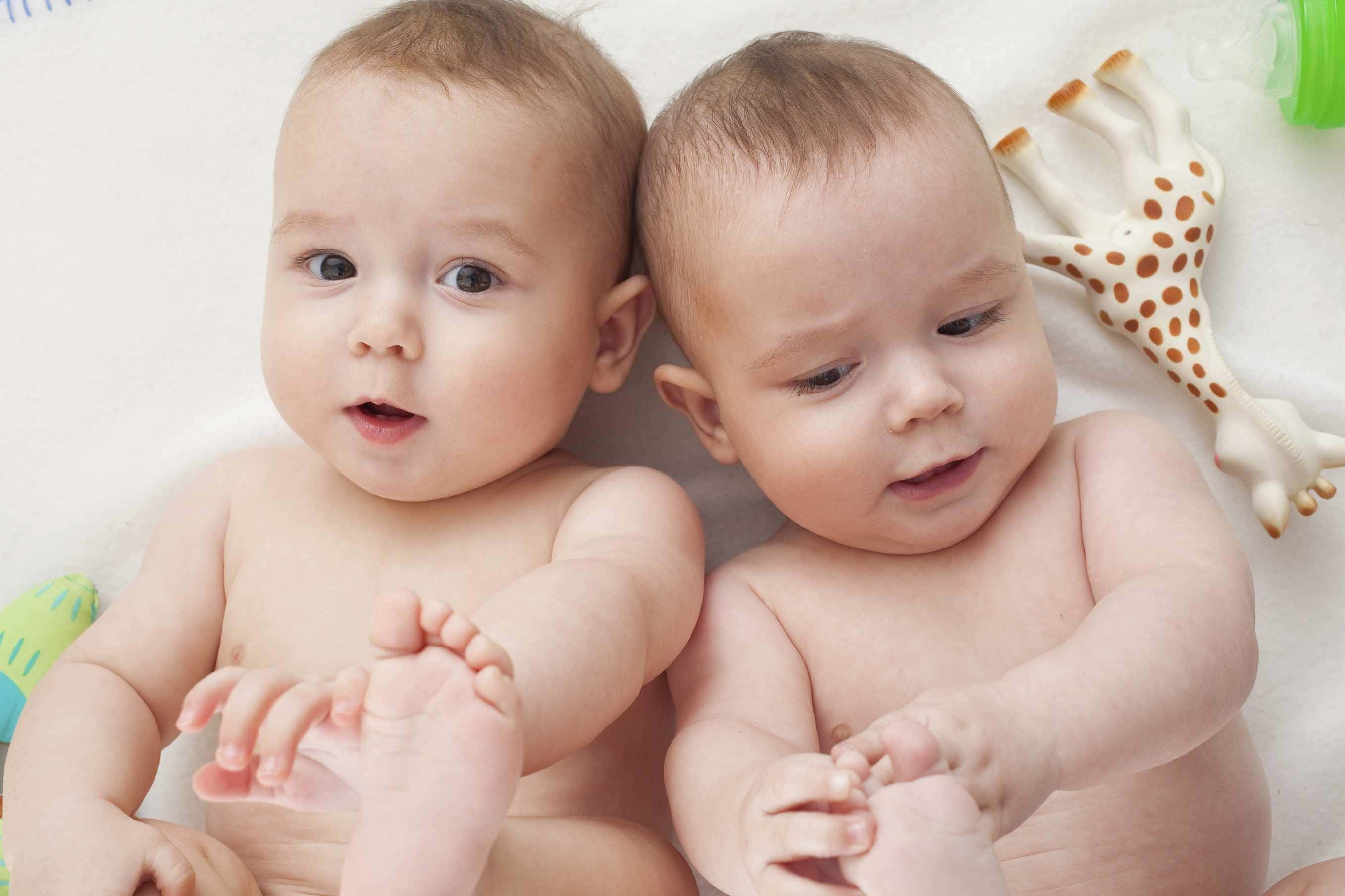 Cute Babies 桌布 壁紙 — 兩個好奇的新生雙胞胎並排躺在柔軟的白色毛毯上，附近有一個長頸鹿牙膠。