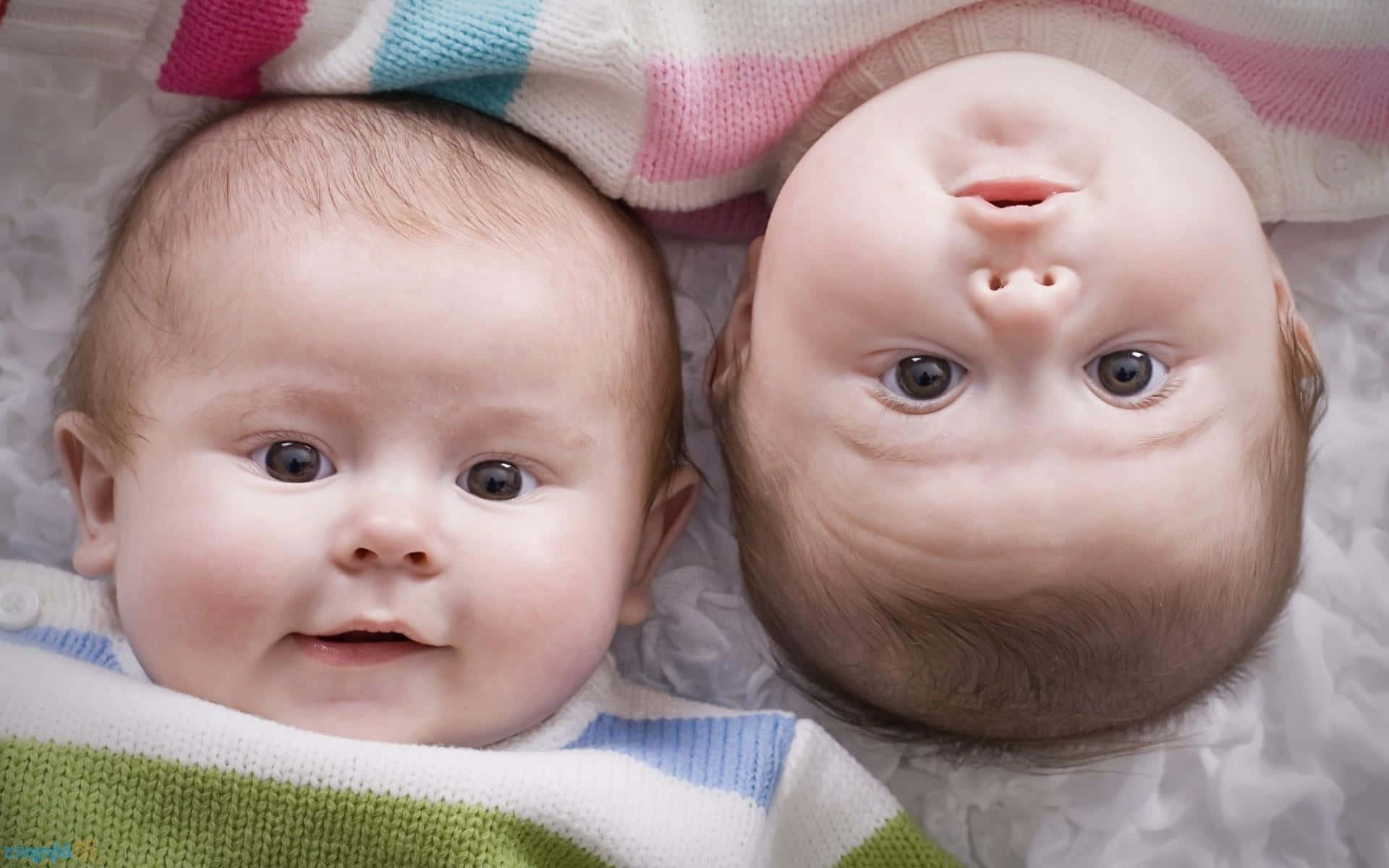 twins 桌布 壁紙 — 兩個嬰兒並排躺在柔軟的毯子上，一個自然地微笑，另一個嘟起嘴巴，睜大眼睛。