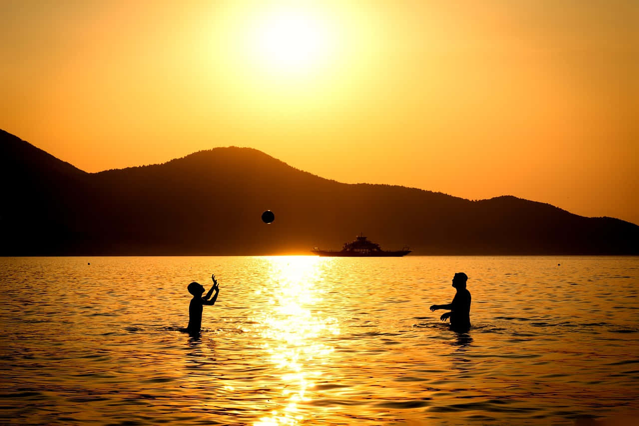 Yellow Beach 桌布 壁紙 — 兩個孩子在金色夕陽的水中玩接球遊戲，背景有山脈剪影和遠處在陽光下閃耀的小船。