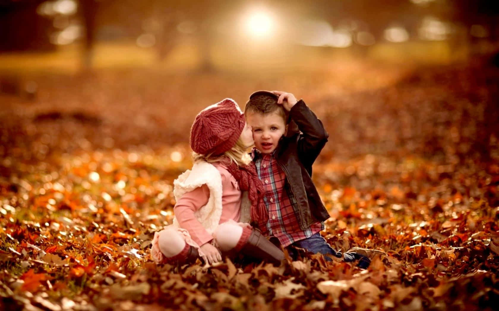 Cute Kids 桌布 壁紙 — 兩個孩子坐在金色秋葉鋪成的地毯中，溫暖的陽光從他們身後的樹木間灑落。