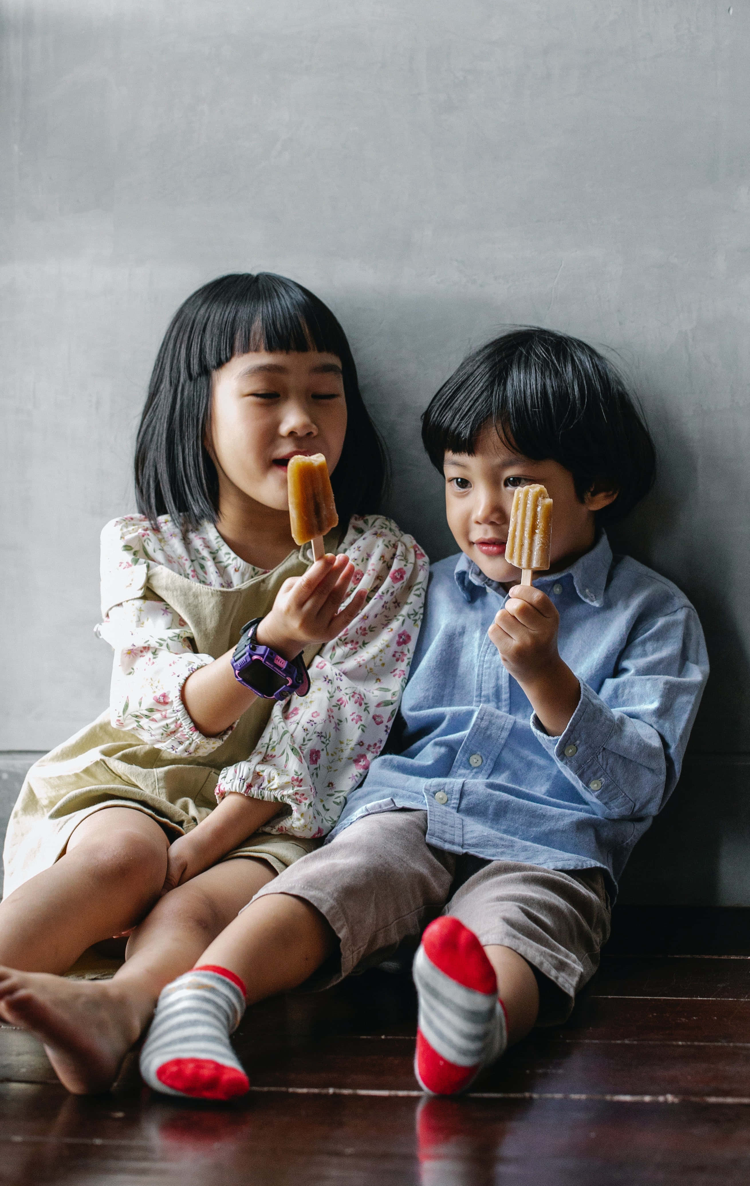 children 桌布 壁紙 — 兩個孩子赤腳坐在木地板上，背靠灰色牆壁，開心享用冰棒。