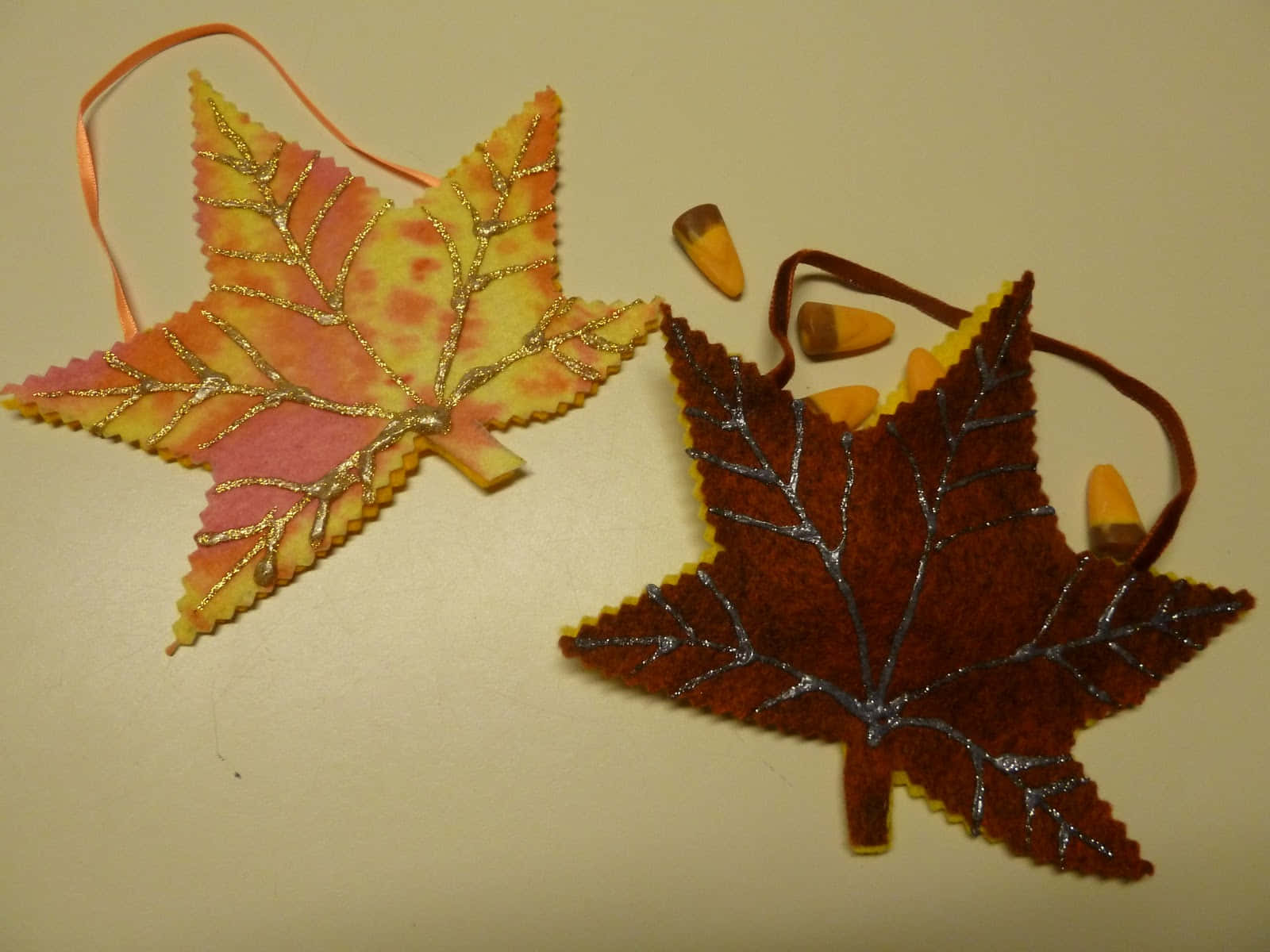 Fall Crafts 桌布 壁紙 — 兩個帶有閃粉葉脈和絲帶環的氈製楓葉裝飾品，放在細小粟米旁邊的純米色表面上。