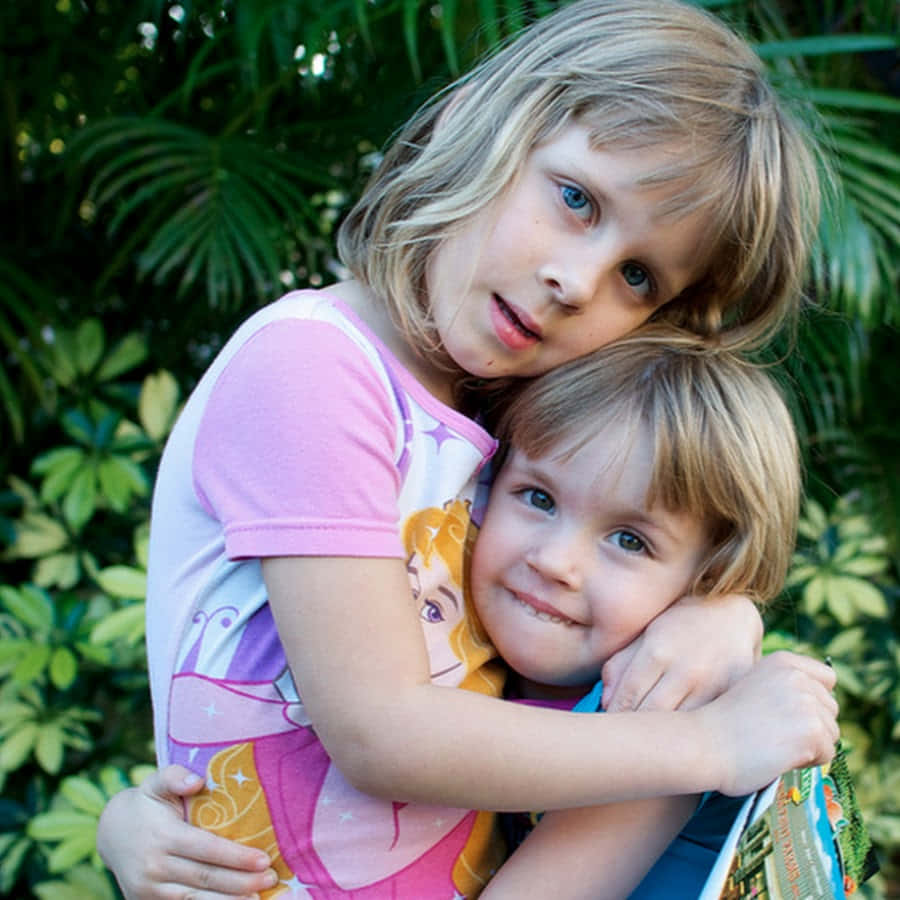 家姐 桌布 壁紙 — 兩個年幼的孩子在陽光明媚的花園裡熱情擁抱，身後是茂密的綠色樹葉。