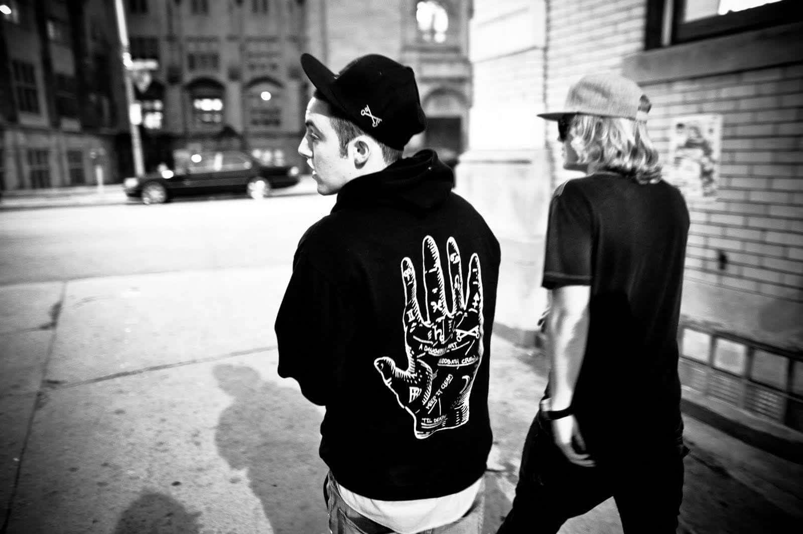 Mac Miller 桌布 壁紙 — 兩個年輕男子夜晚喺城市街道上走開，其中一個著住一件背面有詳細手部圖案嘅連帽衫。