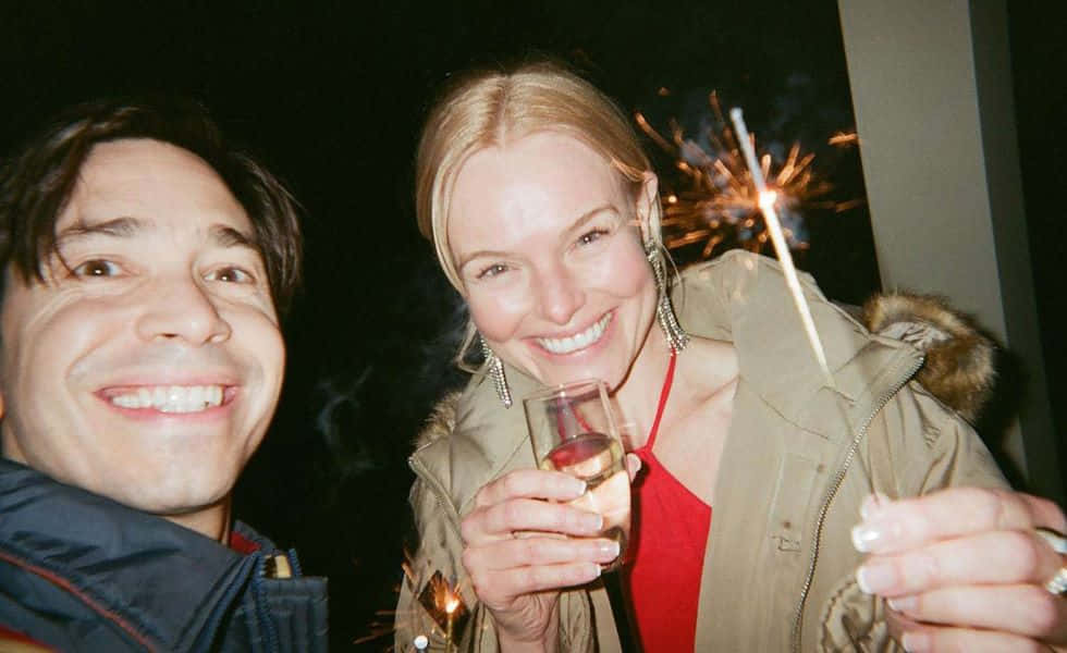 Justin Long 桌布 壁紙 — 兩個微笑的人在夜間戶外節日環境中手持煙花棒和香檳慶祝。