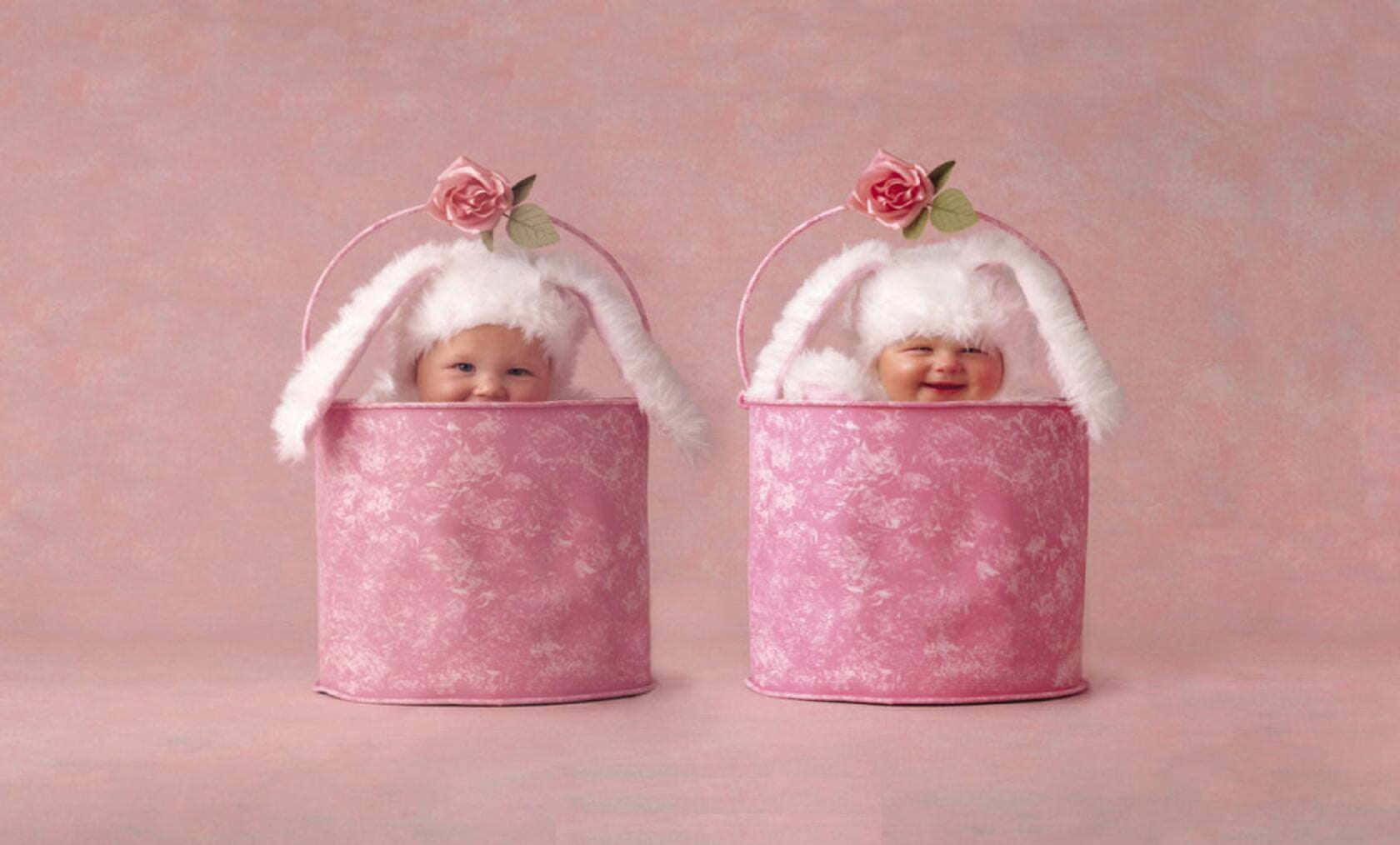 Cute Babies 桌布 壁紙 — 兩個微笑的嬰兒戴著粉紅色兔仔帽，從相配的花卉圖案桶中探出頭來，背景是柔和的粉紅色。