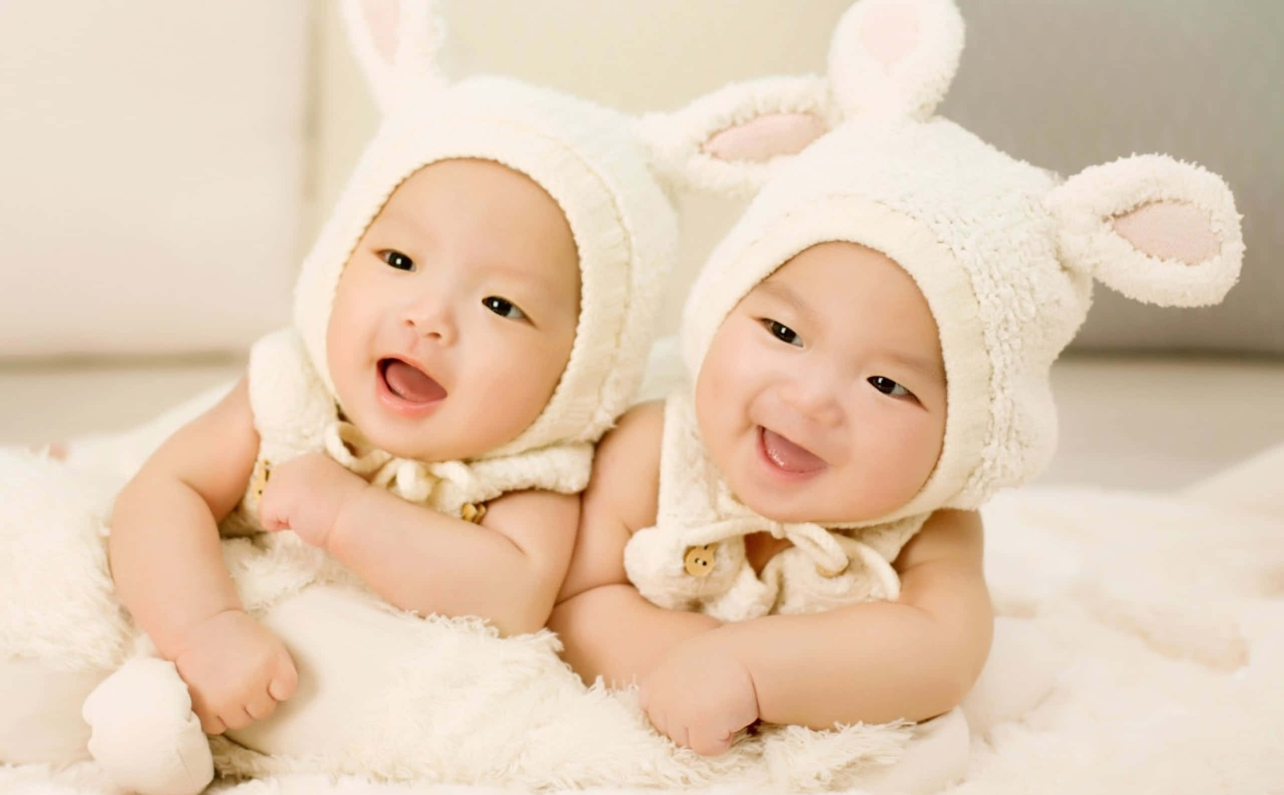 可愛微笑嬰兒 壁紙 — 兩個快樂的嬰兒戴著毛茸茸的白色兔帽，坐在柔軟的白色地毯上開懷大笑。