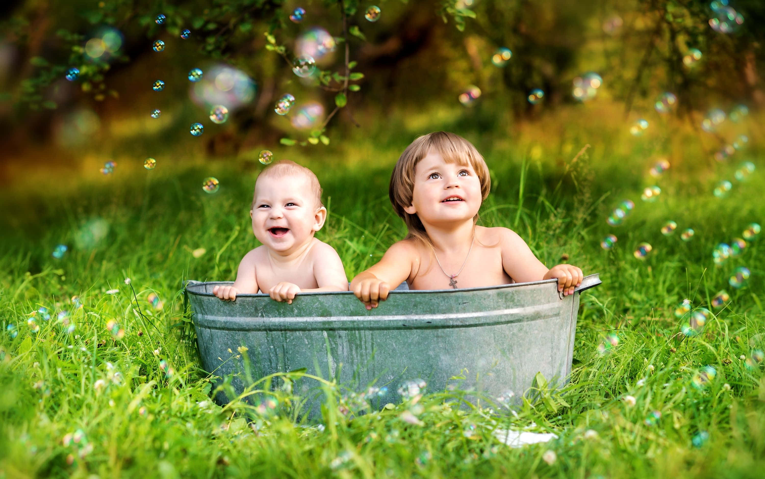 Cute Kids 桌布 壁紙 — 兩個快樂的幼兒在溫暖陽光下，於充滿泡泡的金屬浴盆中嬉笑玩耍，四周是翠綠的草地。