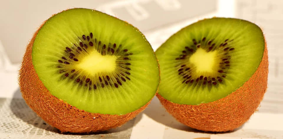 Winter Fruits 桌布 壁紙 — 兩個成熟的奇異果切開一半，露出鮮綠色的果肉和黑色的種子，放在有紋理的表面上。