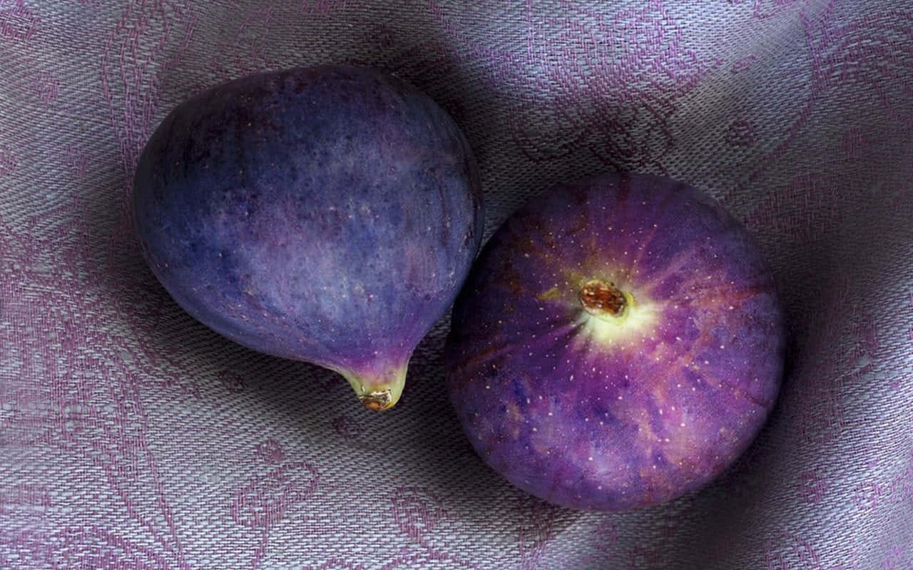 Purple Figs 桌布 壁紙 — 兩個成熟的紫色無花果放在有紋理的薰衣草色布料上，帶有柔和陰影和自然高光。