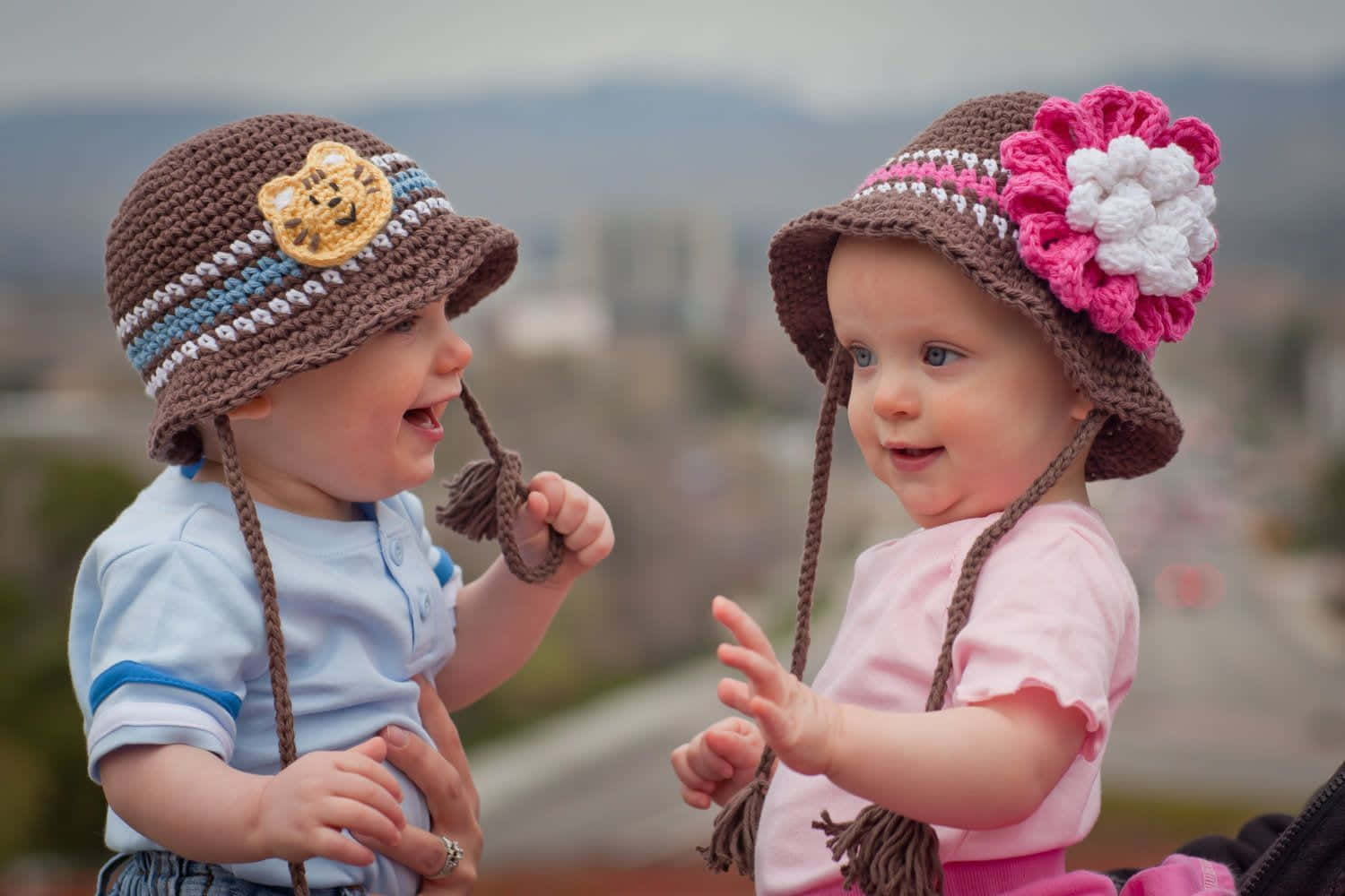 Cute Babies 桌布 壁紙 — 兩個戴著鉤針帽子的微笑幼兒在模糊的城市景觀背景下嬉戲互動。