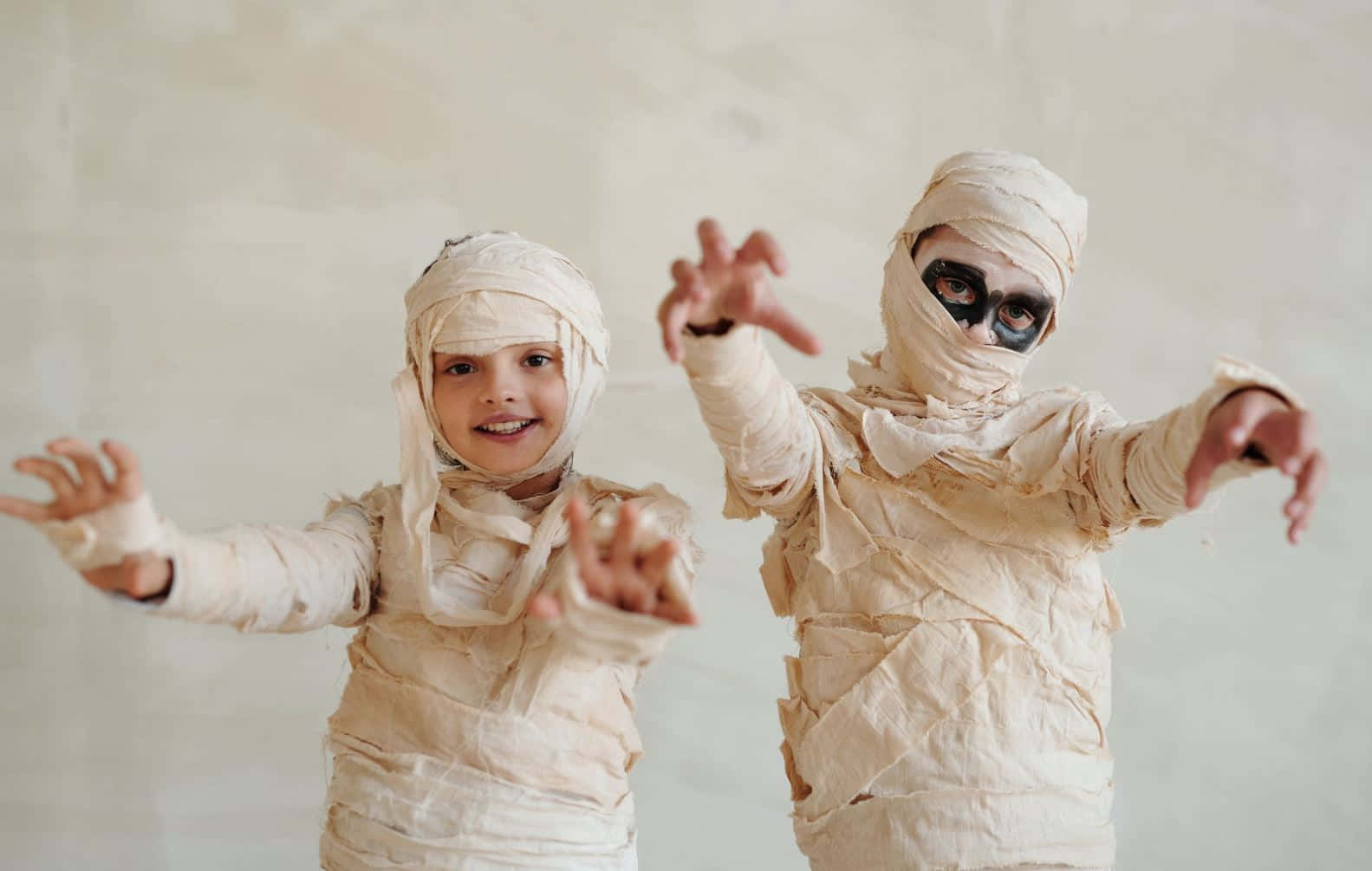 Halloween Costumes 桌布 壁紙 — 兩個打扮成木乃伊的小孩在純米色牆前頑皮地張開雙臂擺姿勢。