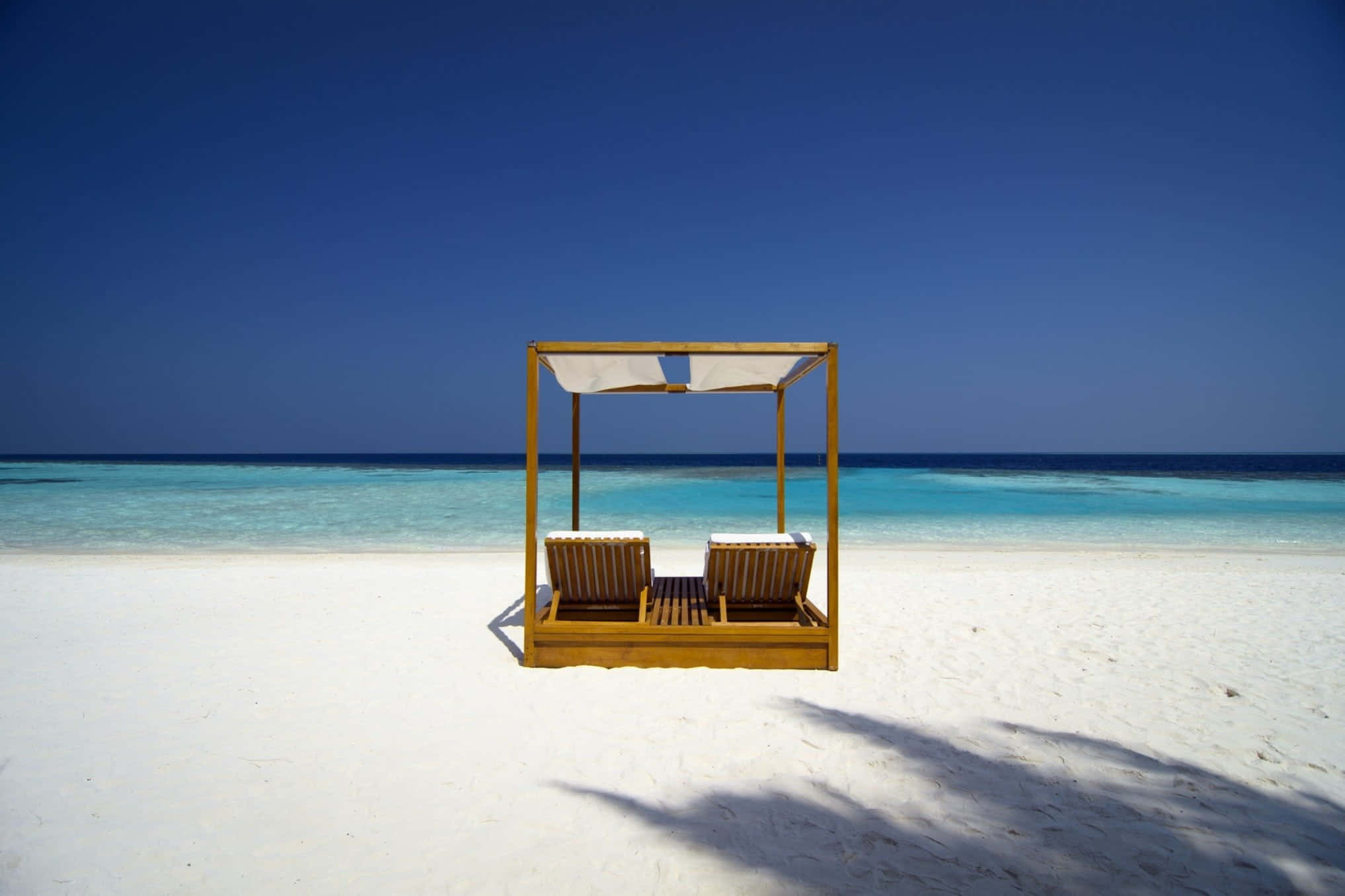 Beach Daybed 桌布 壁紙 — 兩個木製躺椅在遮陽篷下，位於純淨白沙灘上，面向湛藍海洋，天空清澈蔚藍。