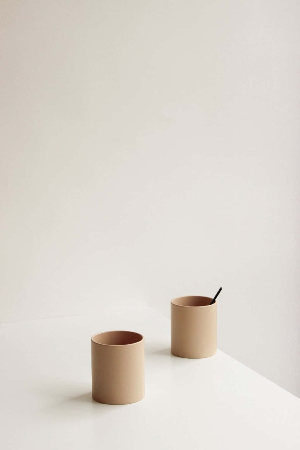 Minimalist Photography 桌布 壁紙 — 兩個極簡風格的米色陶瓷杯放在白色表面上，其中一個拿著黑色湯匙，背景是純淨的米白色牆壁。