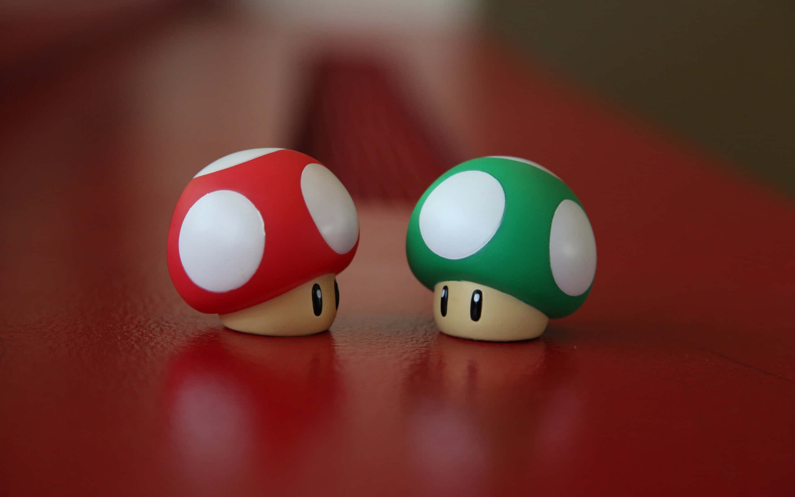 Kawaii Mushroom 桌布 壁紙 — 兩個標誌性的紅色和綠色蘑菇公仔並排坐在光滑的紅色表面上，背景柔和散景。