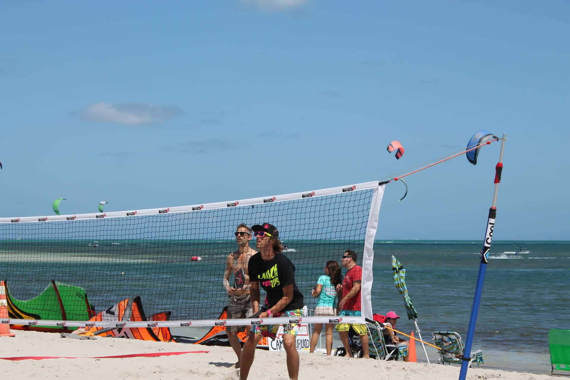 Beach Tennis 桌布 壁紙 — 兩個沙灘排球運動員在陽光明媚的日子裡，附近有風箏衝浪裝備和觀眾，正在比賽中。