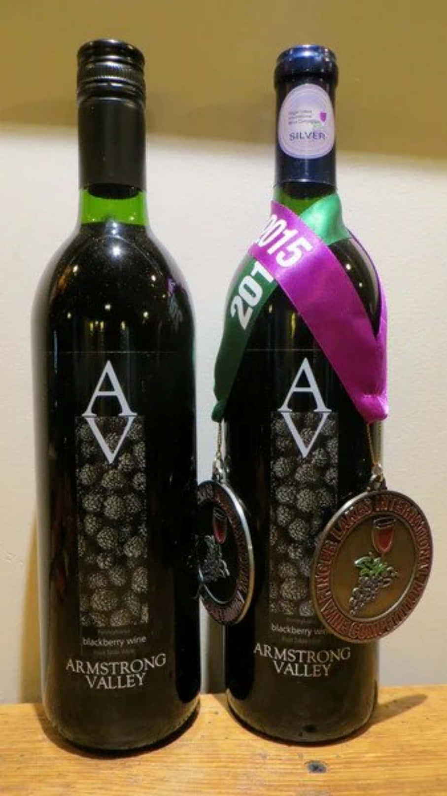 Blackberry Wine 桌布 壁紙 — 兩個深色酒瓶帶有銀色獎牌和紫色絲帶，並排放在木質表面上。