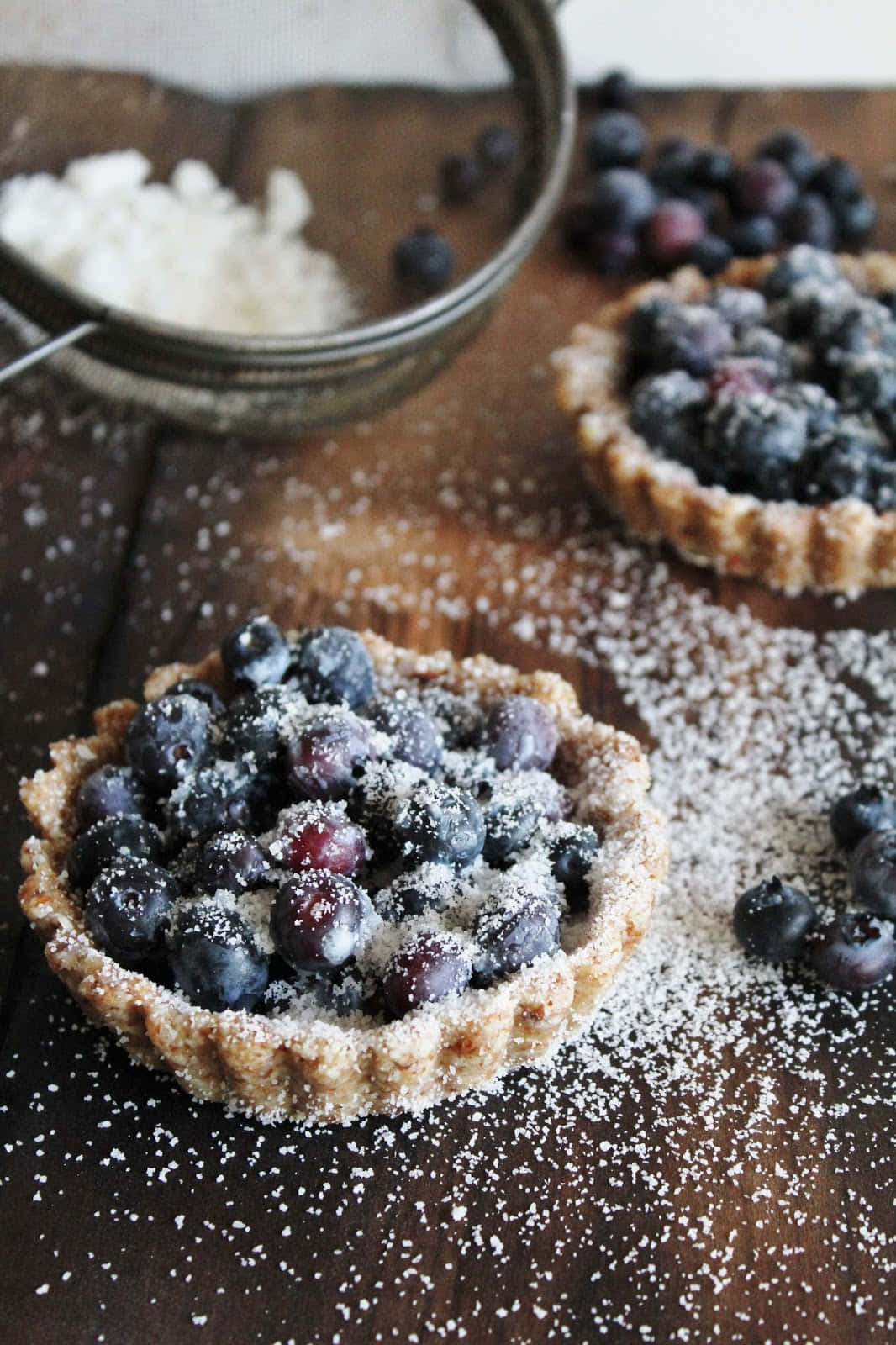Blueberries Tart 桌布 壁紙 — 兩個灑滿糖霜的鄉村藍莓撻放在深色木桌上，旁邊有篩過的糖粉和新鮮莓果。