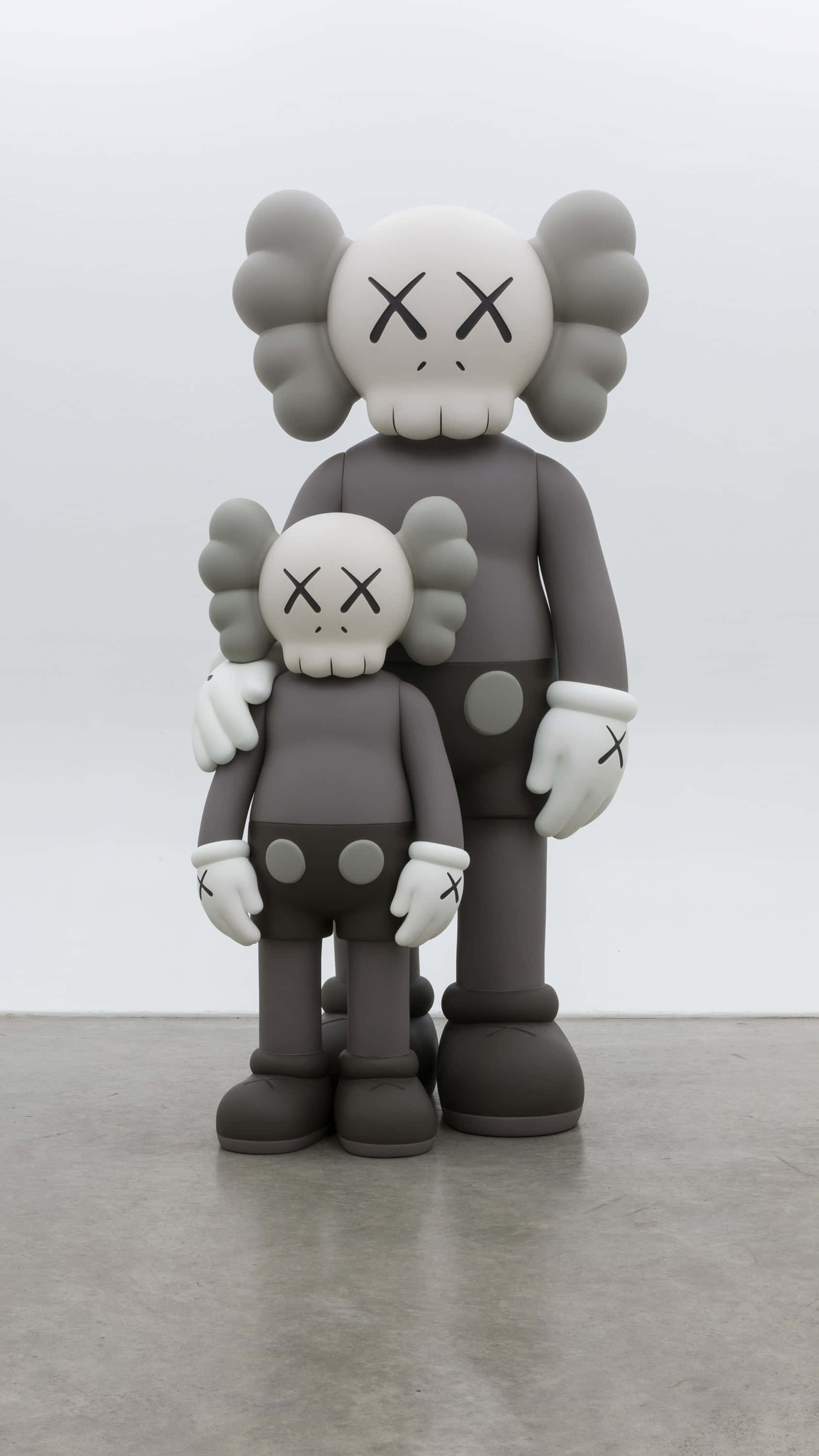 Kaws Bff 桌布 壁紙 — 兩個灰色KAWS Companion公仔，一大一小，並排企喺一個簡約嘅畫廊空間，地板係石屎地，牆壁係白色。